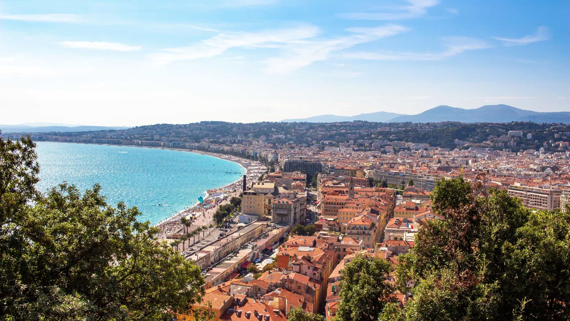 A high angle, panoramic view of the red and orange rooftops of the old town of Nice, France, as seen from the Colline du Château (Castle Hill). The city stretches out below, with the Mediterranean Sea and the Baie des Anges visible in the distance.