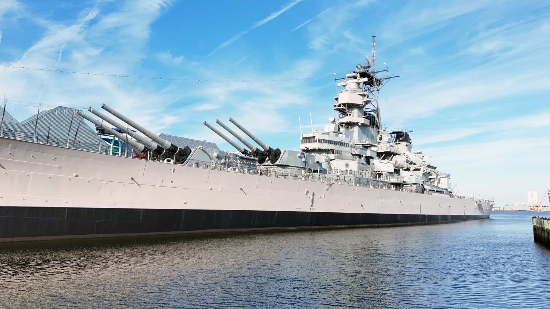 A panoramic view of the massive historic battleship USS Wisconsin (BB-64), an Iowa-class vessel, permanently docked as a museum ship in Norfolk, Virginia. The ship is shown from a distance, displaying its long gray hull, towering superstructure, and the prominent triple-gun turrets. The vessel is tied up at the quayside with a city skyline or port infrastructure visible in the background.