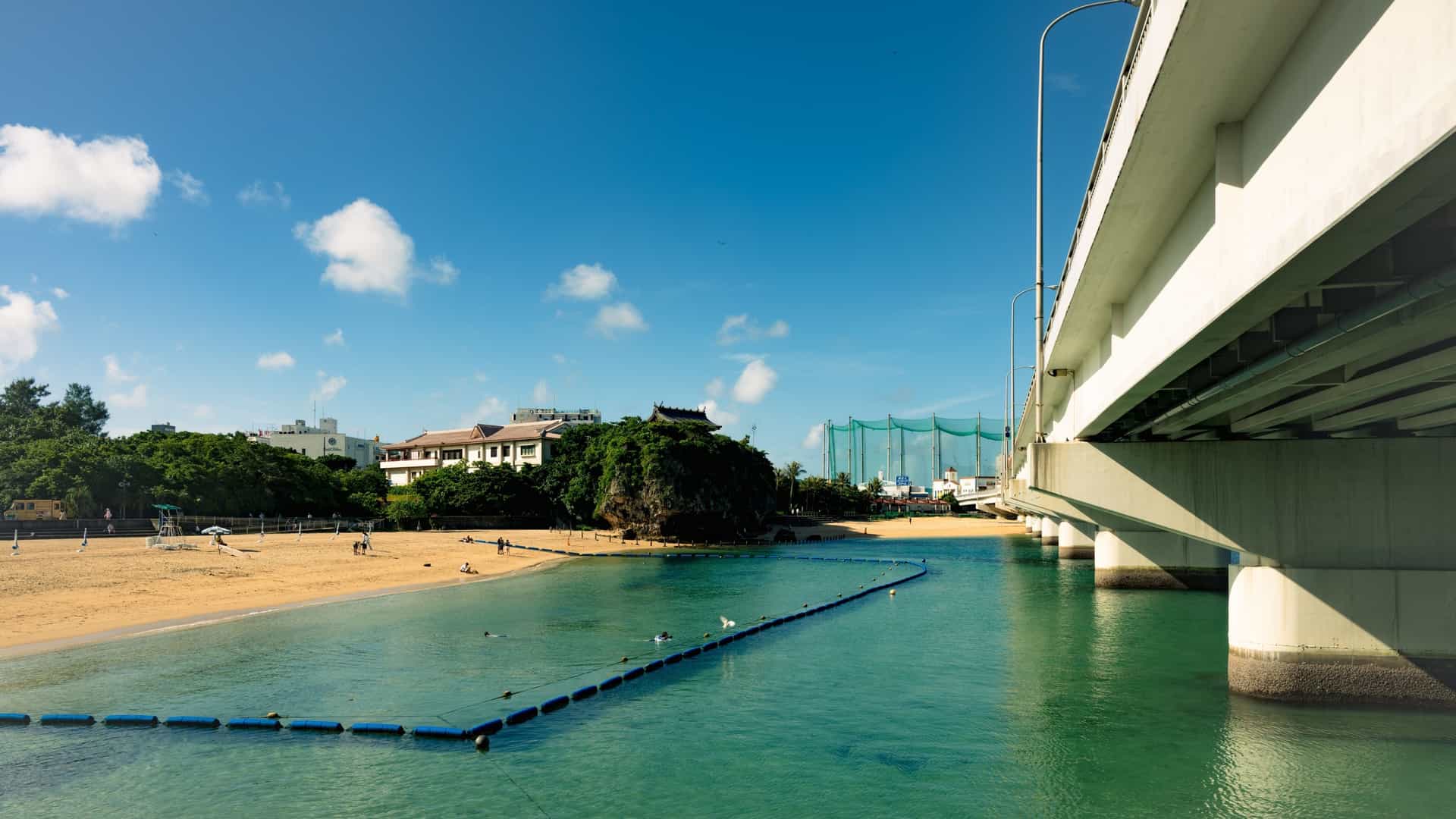 A sunny photograph of Naminoue Beach in Naha, Okinawa, Japan, showing a small, crescent-shaped strip of white sand and turquoise water. The beach is uniquely situated beneath a bridge or elevated highway, with the prominent, distinctive red and white structure of the Naminoue-gū Shrine visible on a cliff overlooking the ocean.