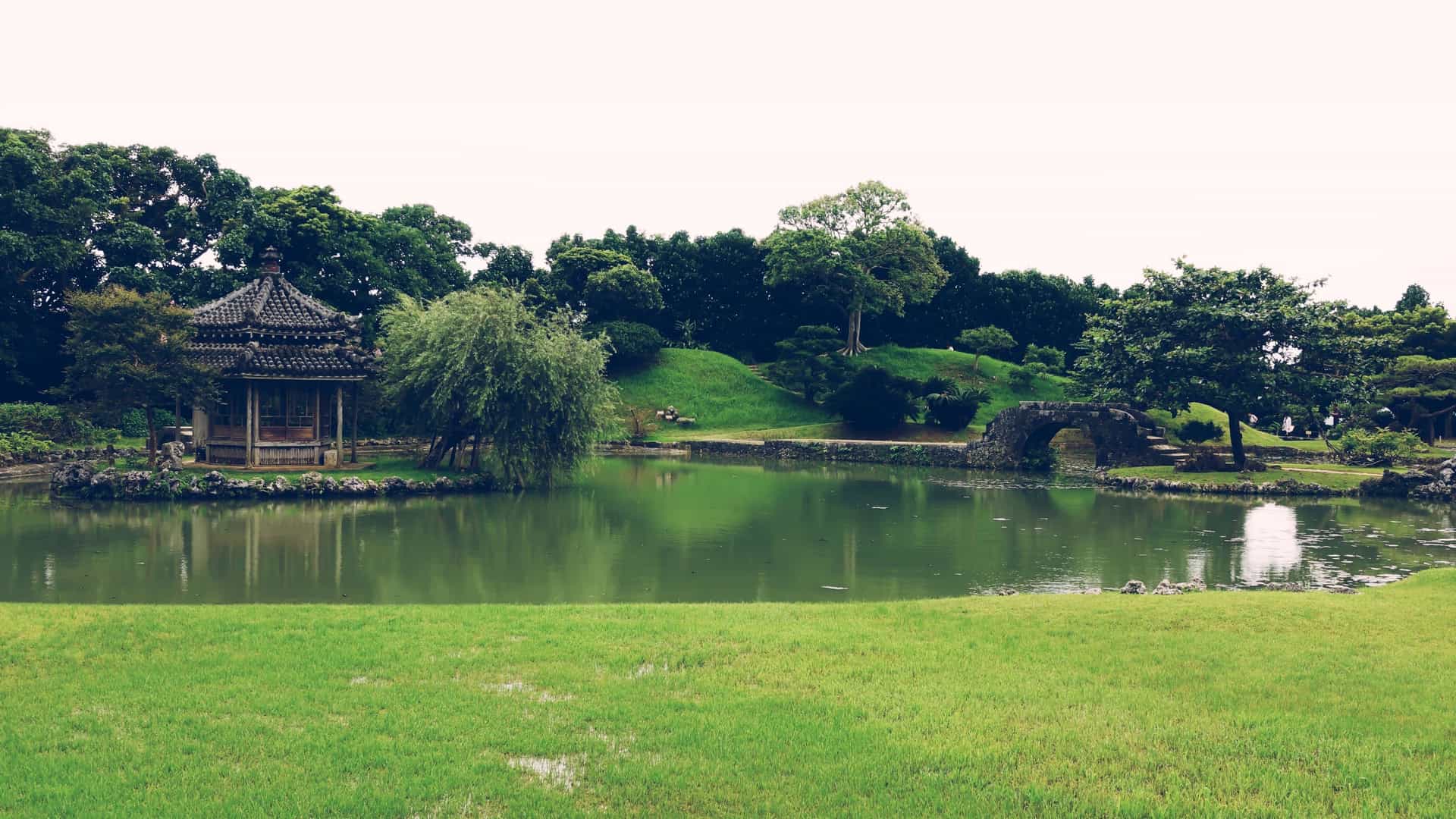 A serene view of the Shikina-en Royal Garden in Naha, Okinawa, Japan, designed in the Ryukyu court style. The image features a large central pond with a wooden, Chinese-style arched bridge, surrounded by lush tropical Okinawan plants, stone lanterns, and traditional wooden palace structures.