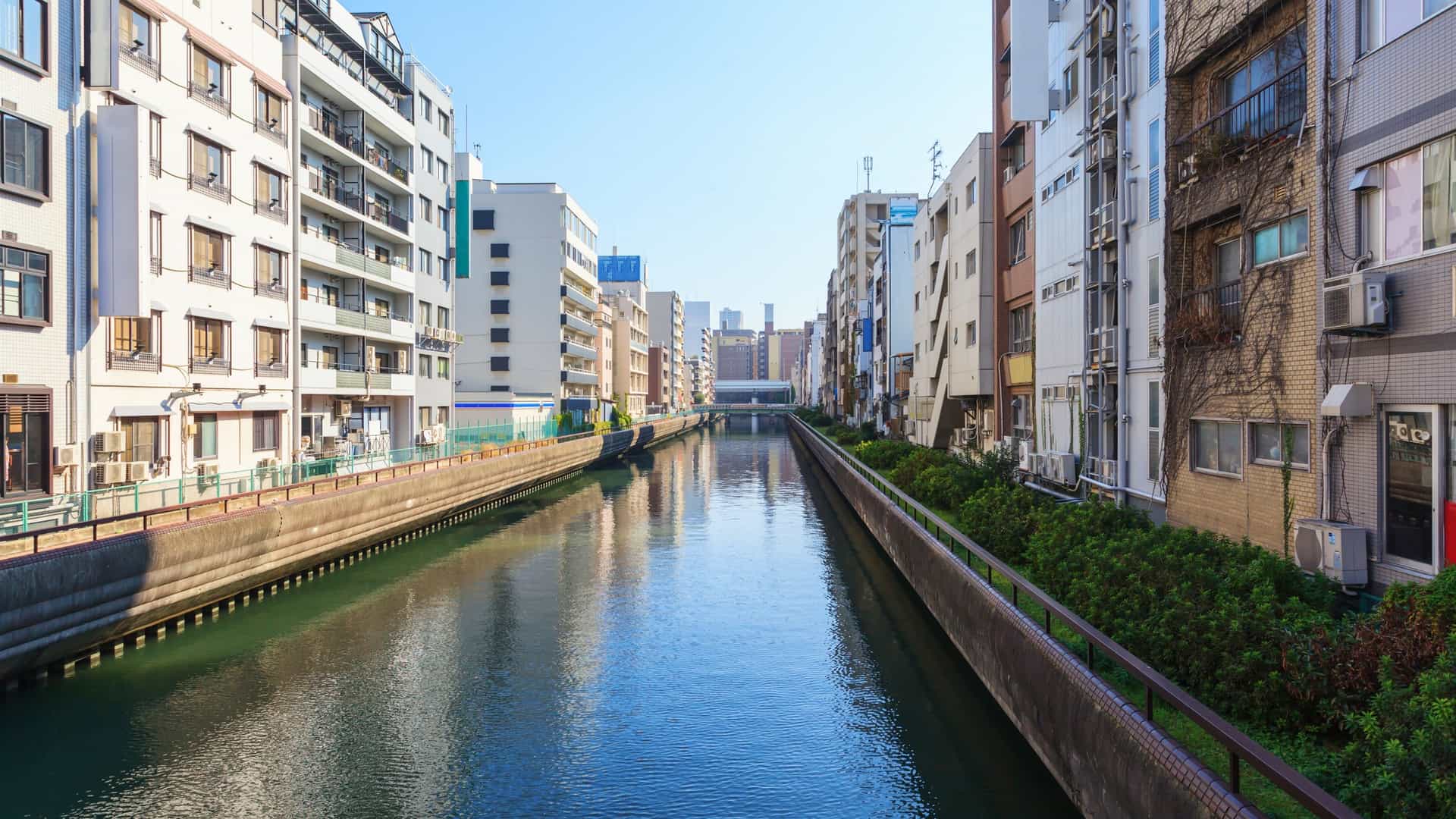 A daytime, eye level view of the Dotonbori canal and walking street in Osaka, Japan. The canal is lined with a variety of colorful buildings, many featuring large, whimsical signs advertising restaurants and shops. The street is bustling with people walking and sightseeing. A boat is visible on the calm water of the canal, and the scene captures the energetic atmosphere of this well known landmark.