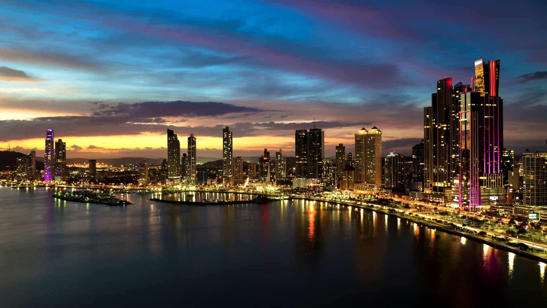 A scenic view of the Panama City skyline and the Cinta Costera, a coastal beltway and park. The modern skyscrapers of the city rise along the waterfront, and a major highway and pedestrian path curve along the bay. A body of water, likely the Pacific Ocean, is visible in the foreground. The image captures the vibrant, urban atmosphere of Panama City's waterfront.