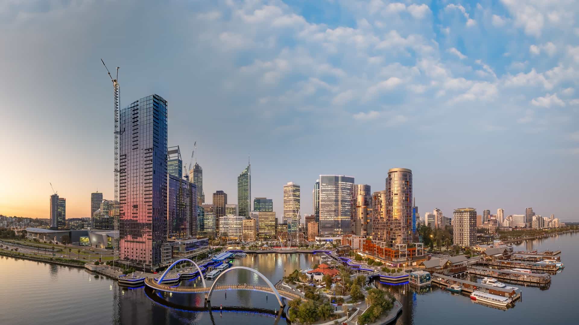 A panoramic sunset view of Elizabeth Quay in Perth, Western Australia. The image shows the modern city skyline reflecting in the tranquil waters of the Swan River, with the Elizabeth Quay Bridge as a prominent feature. The sky is filled with the warm, vibrant colors of the setting sun.