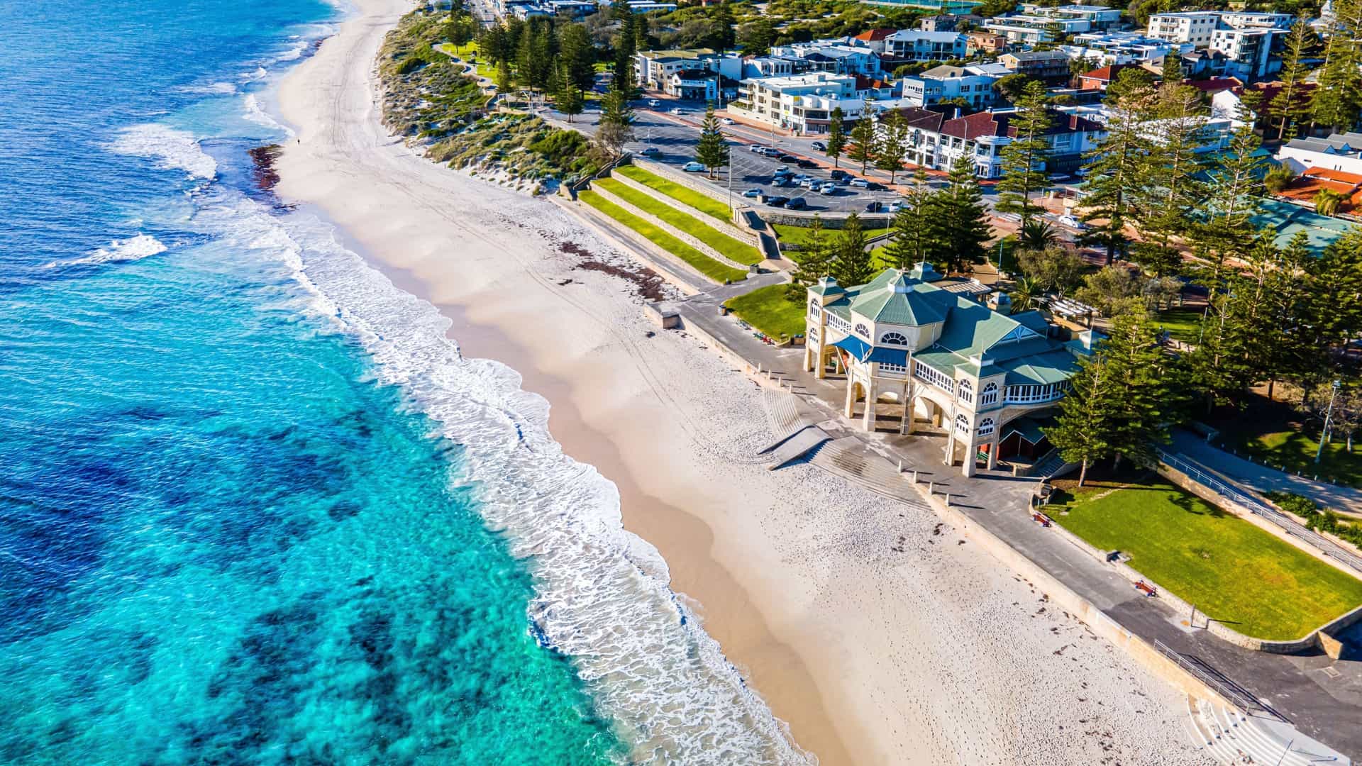 An aerial view of Cottesloe Beach in Western Australia, with stunning crystal blue waters and a white sandy shore. The image also features the iconic Indiana Tea House and a lively coastal atmosphere.
