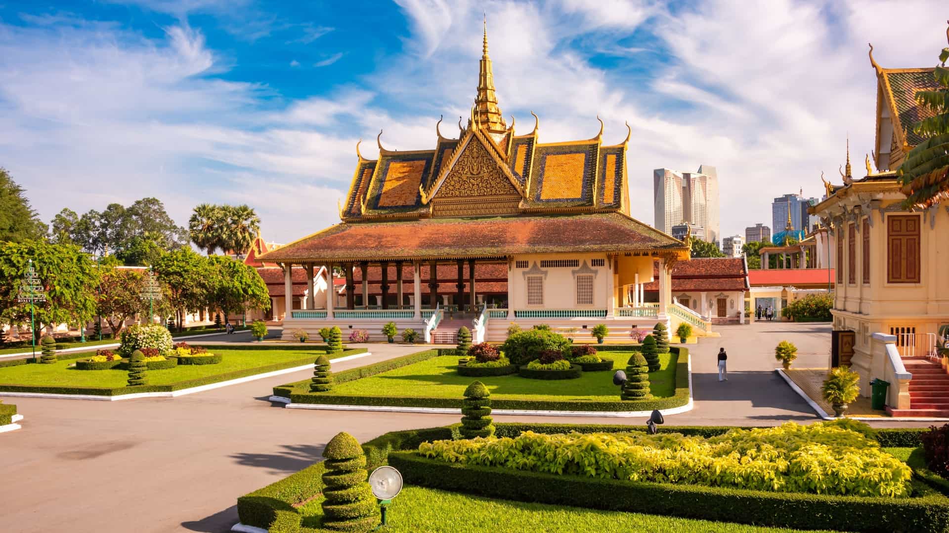 A scenic view of the Royal Palace in Phnom Penh, Cambodia, with its complex of ornate buildings set against a clear blue sky. The image highlights the traditional Khmer architecture, featuring intricate golden roofs and multi tiered spires. In the foreground, there is a large, well maintained lawn, and a wide path leads toward the palace entrance.
