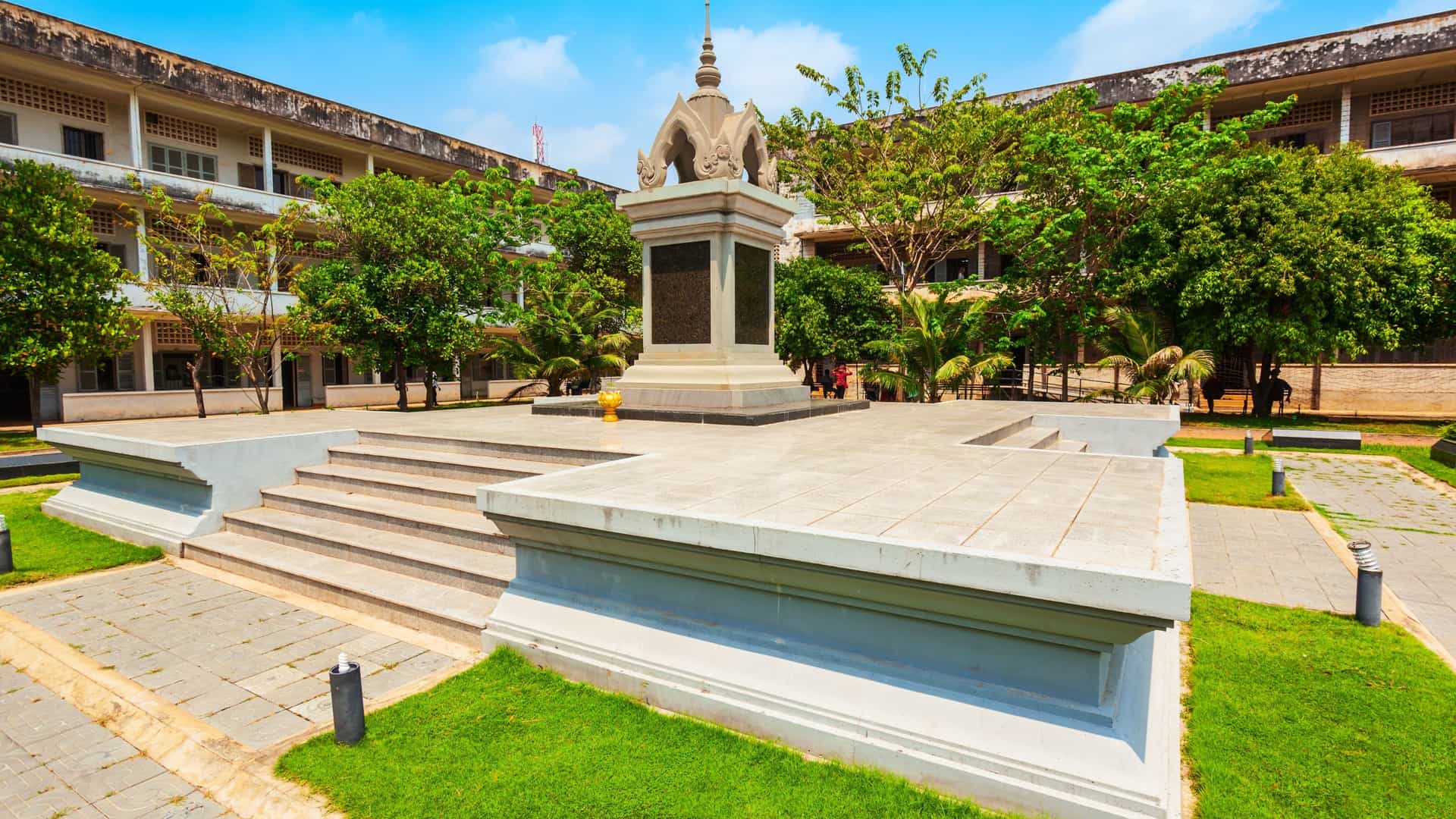 A view of the Tuol Sleng Genocide Museum in Phnom Penh, Cambodia. The building, a former high school, has a stark, institutional appearance with multi story, whitewashed facades, balconies with railings, and barred windows. The image captures the chilling atmosphere of the former S 21 prison, a place of mass incarceration and torture under the Khmer Rouge regime.