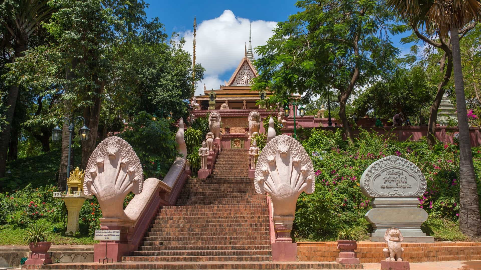 A serene view of Wat Phnom temple, a prominent Buddhist religious site located on a hill in Phnom Penh, Cambodia. The temple building features a traditional Khmer architectural style with a multi tiered, golden roof and a central spire. The building is surrounded by lush green trees, and a stupa is visible to the side.