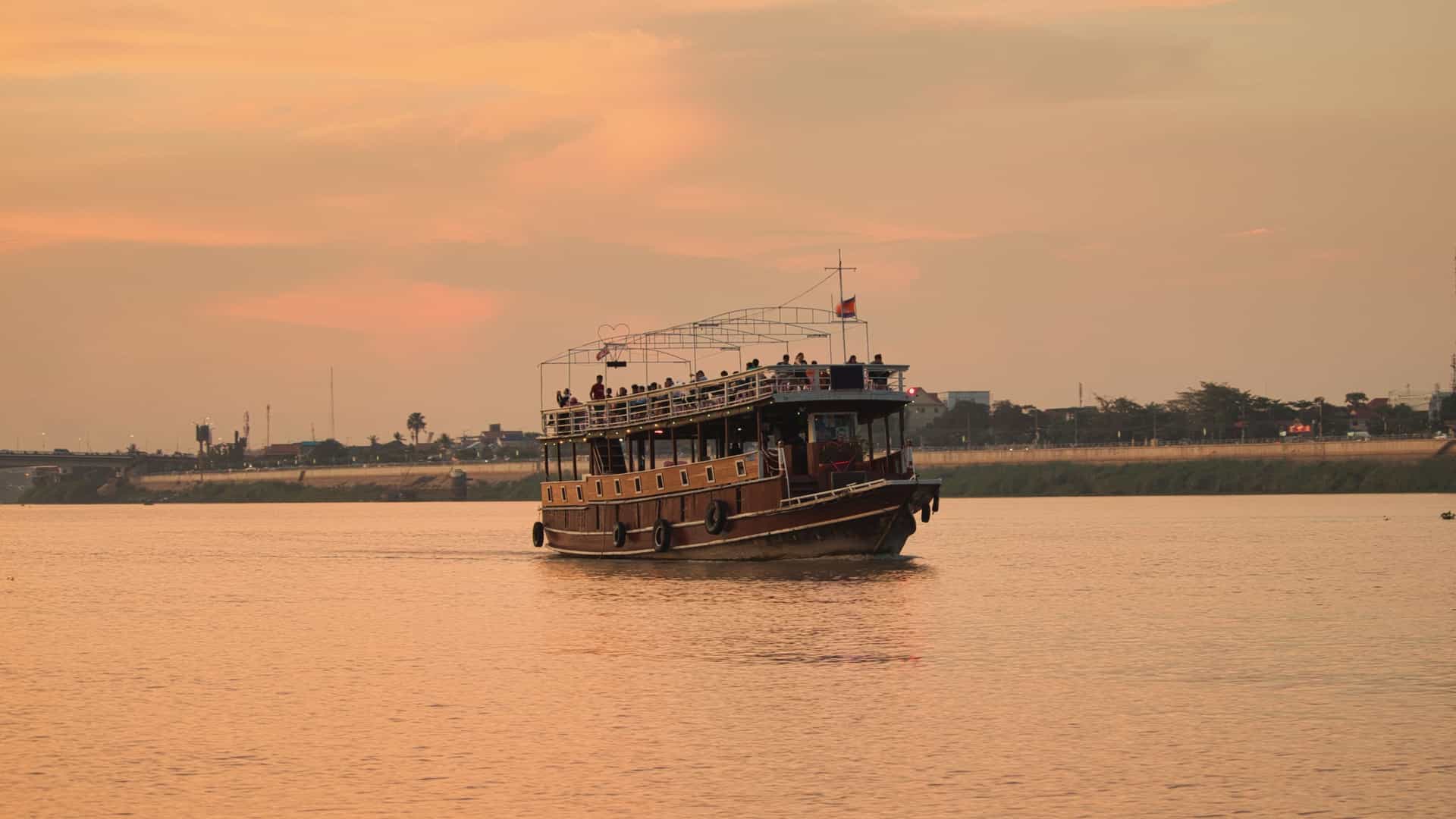 A tourist boat, with an open upper deck, sails on the wide, calm waters of the Mekong River at sunset. The sky is filled with a soft golden light, and the modern cityscape of Phnom Penh is visible in the background, with the developing skyline catching the last rays of the sun.