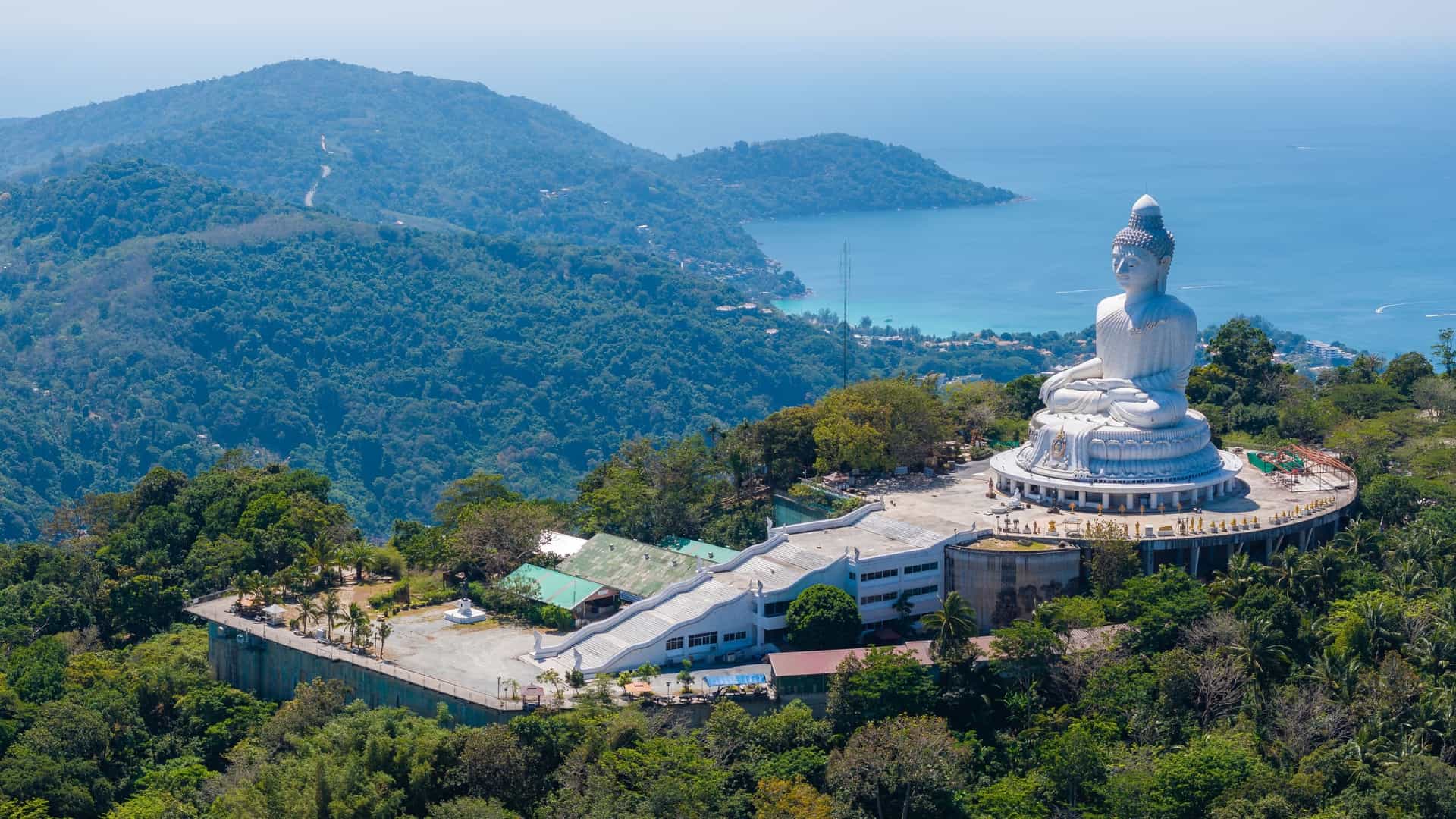 An aerial view of the seated, 45 meter tall Big Buddha statue made of white marble. The statue, located on Nakkerd Hill in Phuket, Thailand, overlooks the lush green landscape and the surrounding bays and coastline, including Chalong Bay.