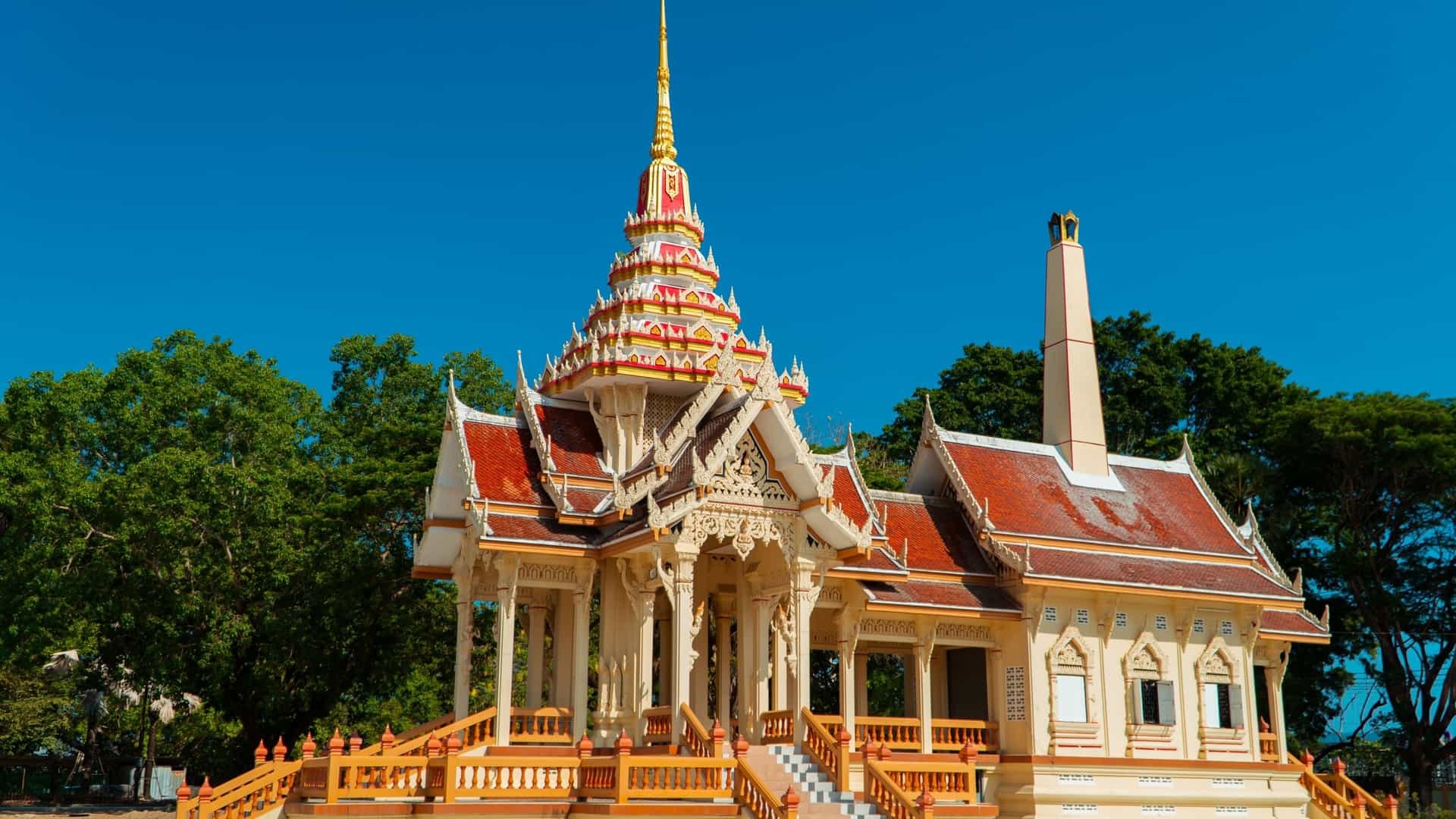 The main chedi, or stupa, of Wat Chalong Buddhist temple in Phuket, Thailand, stands tall with its tiered, pointed golden roof. The ornate building is surrounded by a white wall and smaller, colorful temple structures and statues, with the entire complex set against a clear blue sky.