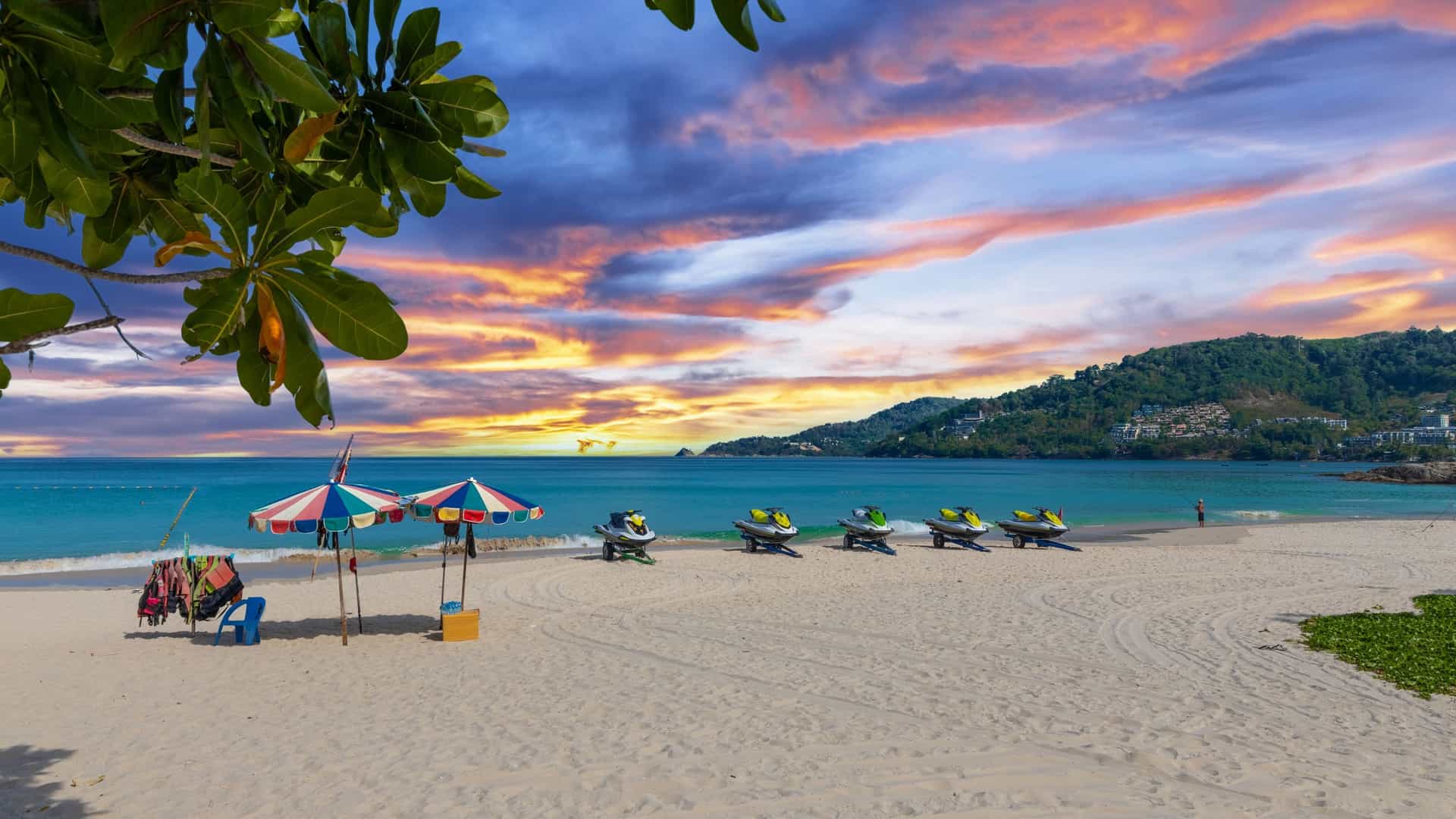 A serene view of Patong Beach in Phuket, Thailand, showing a wide expanse of white sand meeting the calm, turquoise waters of the Andaman Sea. The beach is lined with rows of green palm trees and sun loungers, with a few people scattered along the shore.