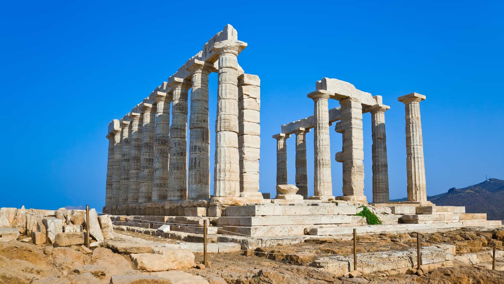 The ancient Temple of Poseidon stands on a rocky cliff at Cape Sounion, Greece. Several tall, weathered Doric columns are visible against the backdrop of the Aegean Sea, with a few small islands on the horizon.