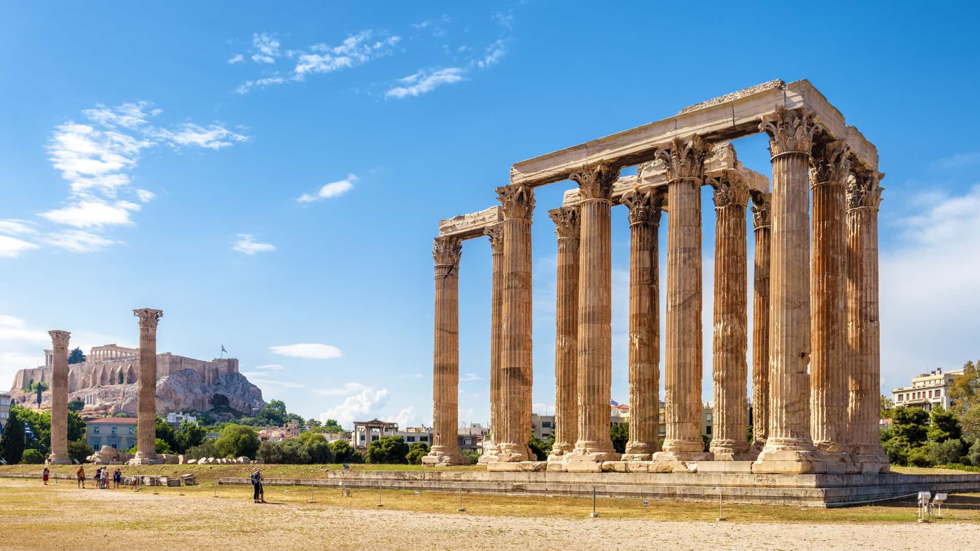 A wide angle view of the Temple of Olympian Zeus in Athens, Greece. The remaining immense Corinthian columns stand in a field of green grass with the ancient Acropolis visible on the hill in the distance under a blue sky.