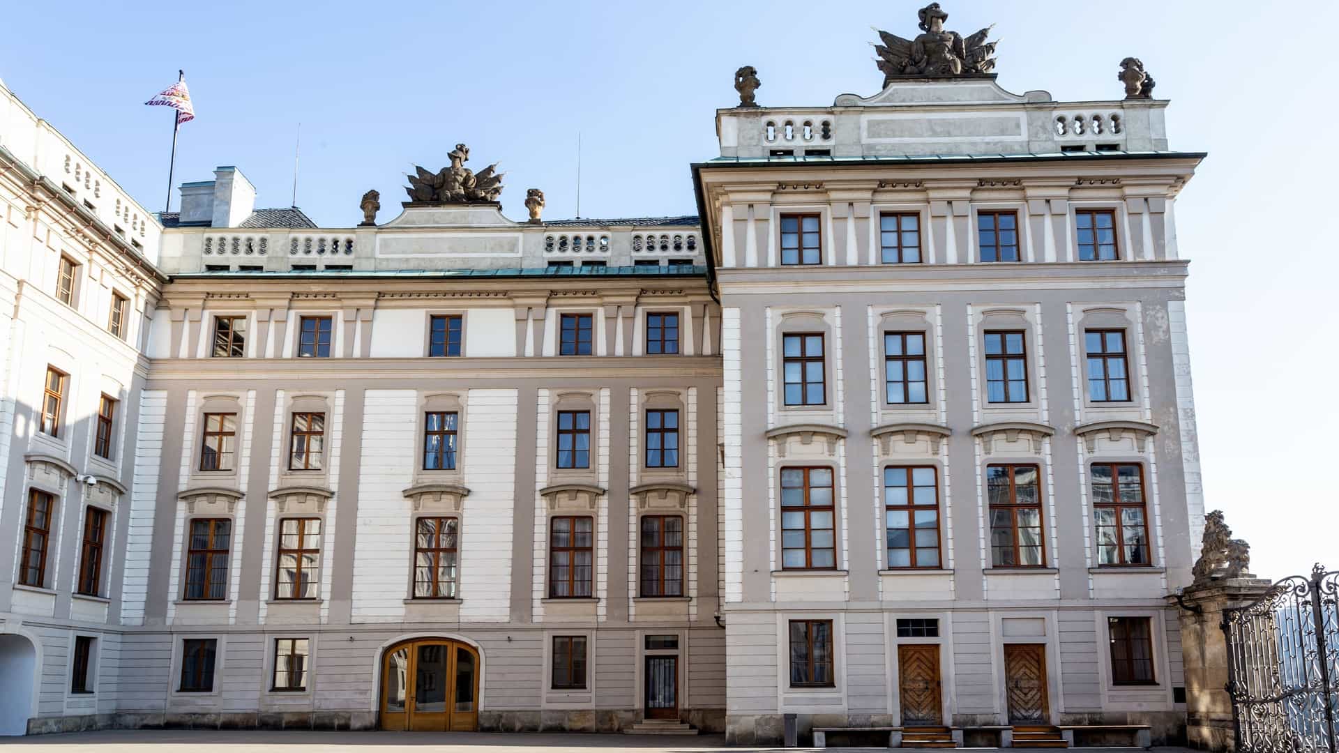 A panoramic view of the Prague Castle complex in the Hradčany district, dominated by the towering Gothic spires of St. Vitus Cathedral, which rises above the surrounding palace buildings and the red tiled rooftops of the historic city below.