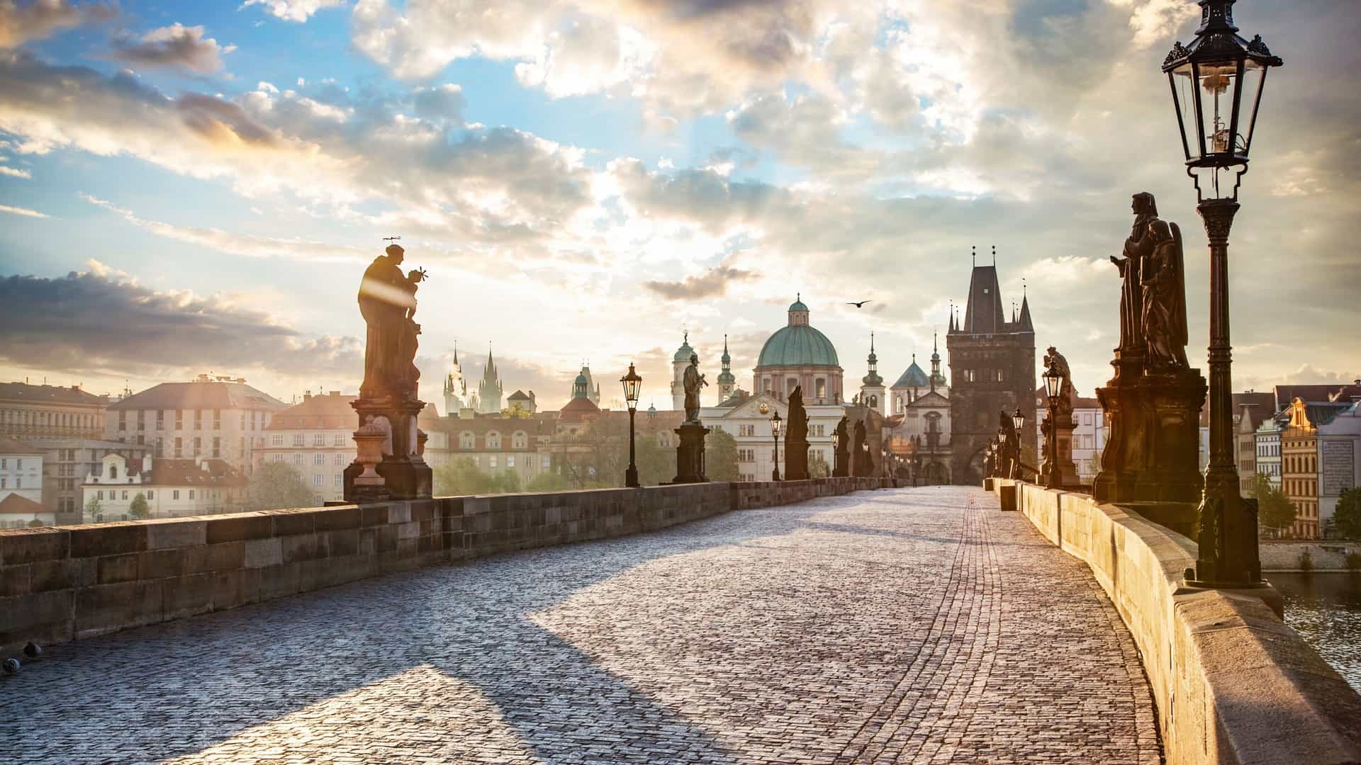 A breathtaking view of the Charles Bridge in Prague at sunrise. The historic stone bridge is lined with Baroque statues and features the silhouettes of people walking across it. In the background, the Old Town Bridge Tower is visible, with the sky glowing with the warm colors of the rising sun.