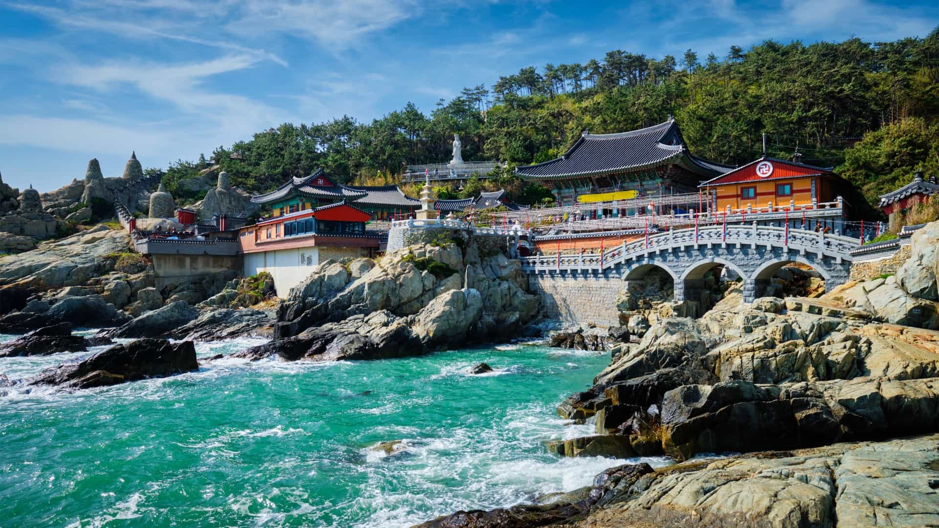 The Haedong Yonggungsa Temple, a traditional Korean Buddhist temple, is shown with its colorful, multi tiered pavilions built directly on a rocky cliff overlooking the blue ocean in Busan, South Korea.