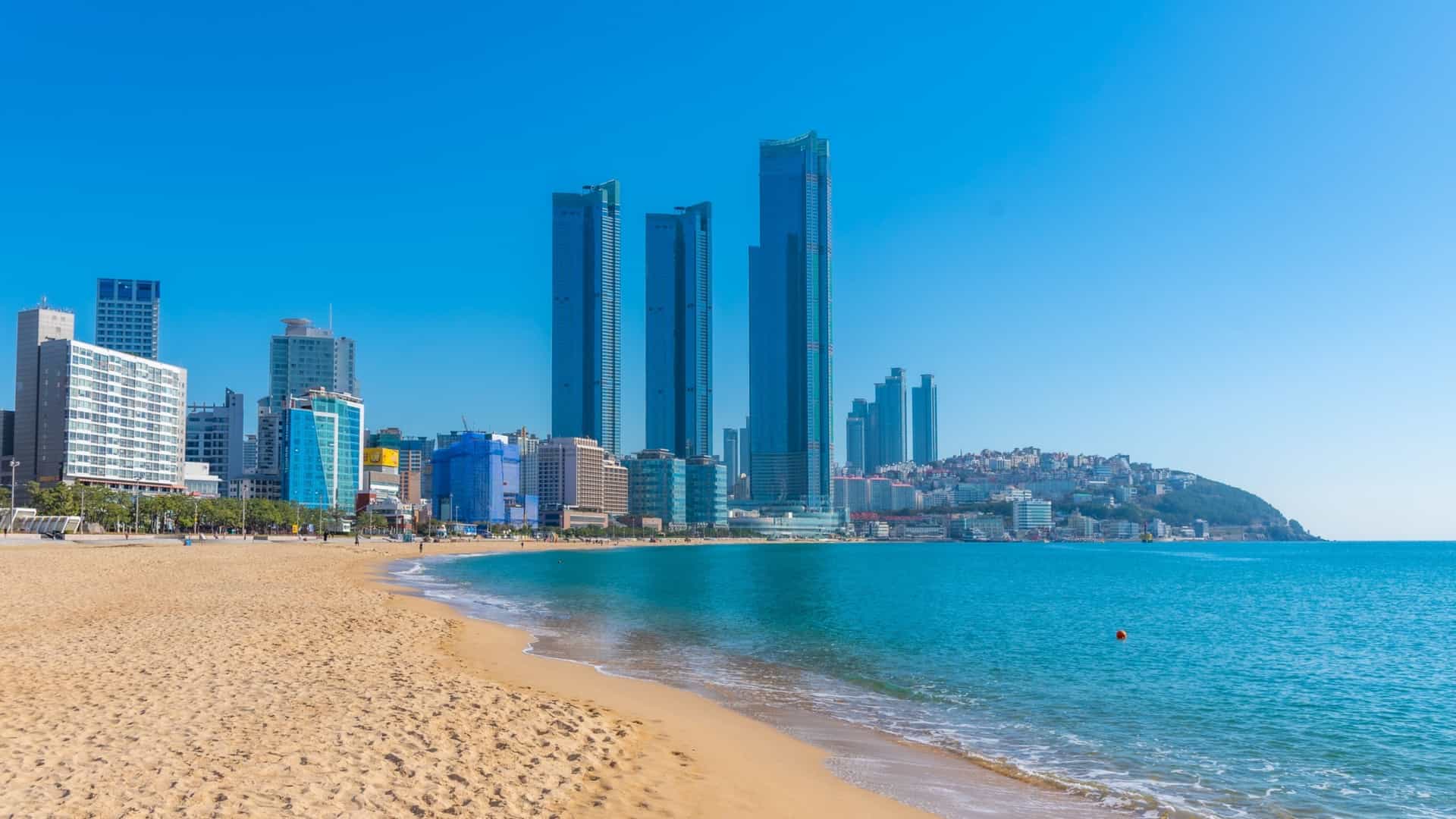 A wide, sandy expanse of Haeundae Beach in Busan, South Korea, with a row of modern skyscrapers and high rise apartments lining the back of the beach, all set against the blue water of the sea and a clear sky.