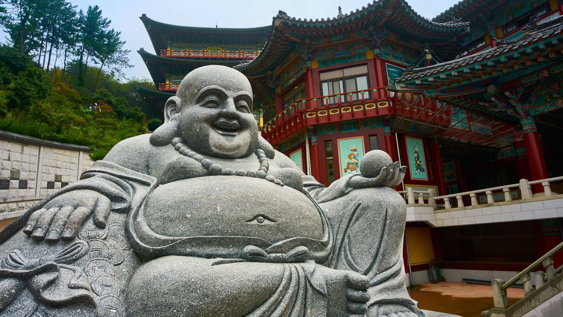 A photograph of the Beomeosa Temple complex, a major Korean Buddhist temple located on the slopes of Geumjeongsan Mountain in Busan, South Korea. The image features traditional Korean architecture with vibrant dancheong (ornate coloring) on the wooden buildings, set against a backdrop of green trees.
