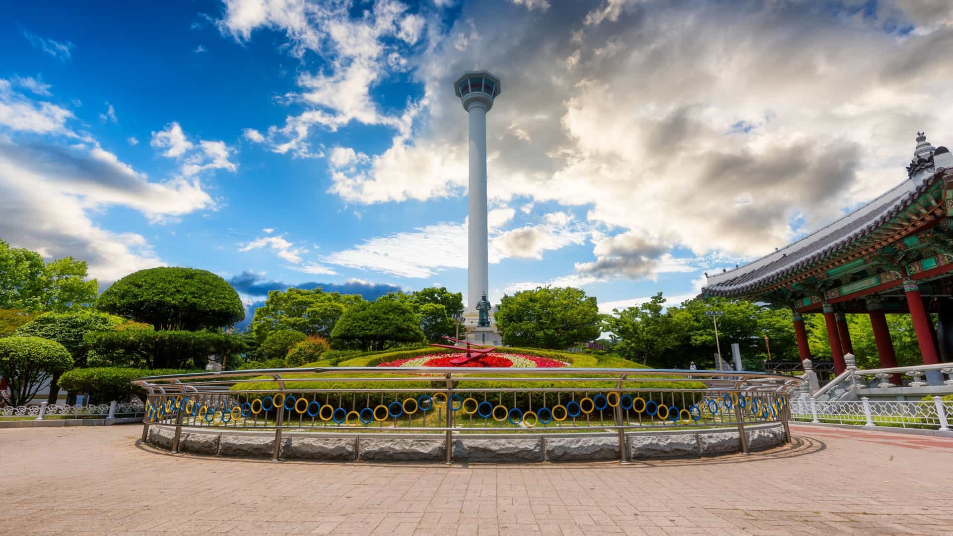 A daytime photograph of the Busan Tower (also called Busan Diamond Tower), a white observation tower in Yongdusan Park, Busan, South Korea. The tall, distinctively shaped tower is a major city landmark, and is surrounded by trees in the park's elevated setting.