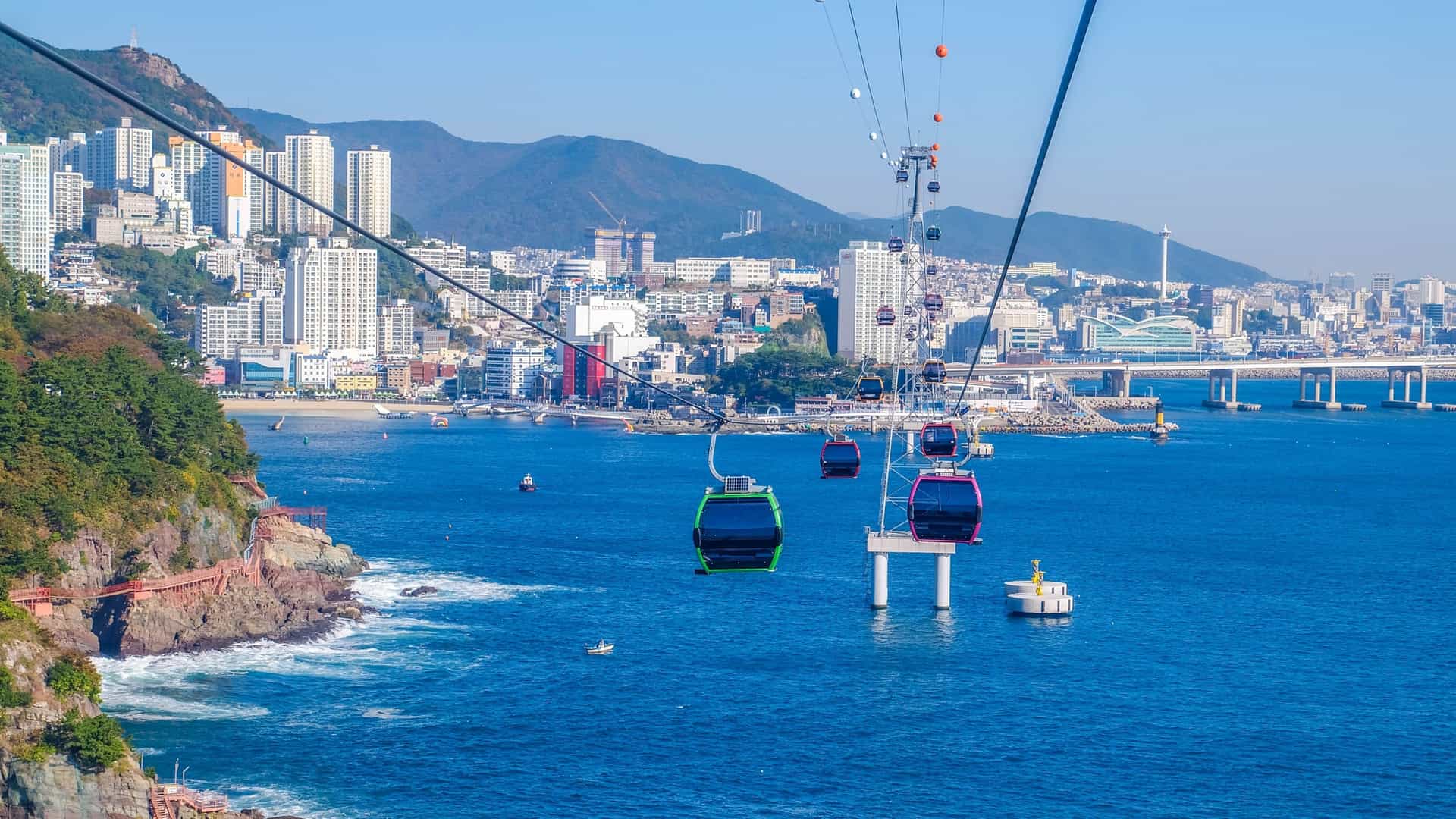 An impressive aerial view showing a red Songdo Marine Cable Car gondola soaring high above the sea in Busan, South Korea. The cable car transports passengers over the bright blue water toward the city's coastal area. In the background, a dense city skyline and a large suspension bridge (the Namhangdaegyo Bridge) are clearly visible, highlighting the scenic urban and marine landscape.