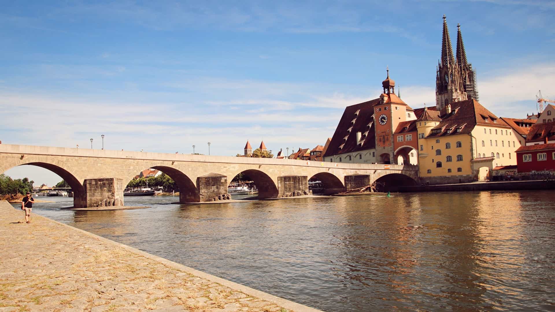The ancient, arched Stone Bridge crosses the Danube River in Regensburg, Germany, with historic Old Town buildings and cathedral towers visible under a clear, sunny sky.