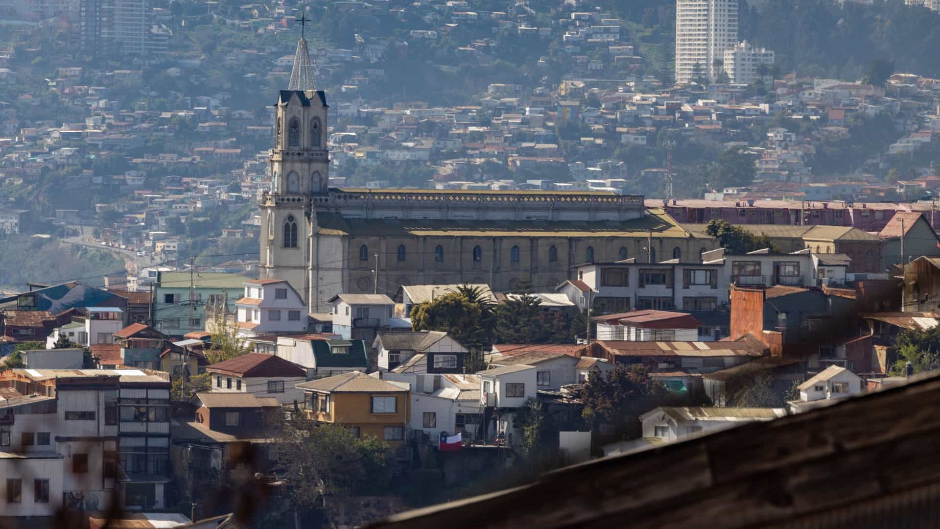 A colorful, panoramic view from Cerro Concepción in Valparaíso, Chile, showcasing the steep hillside densely covered with a vibrant mosaic of historic houses and buildings. The colorful cityscape extends down toward the Pacific Ocean, visible in the background under a wide sky.