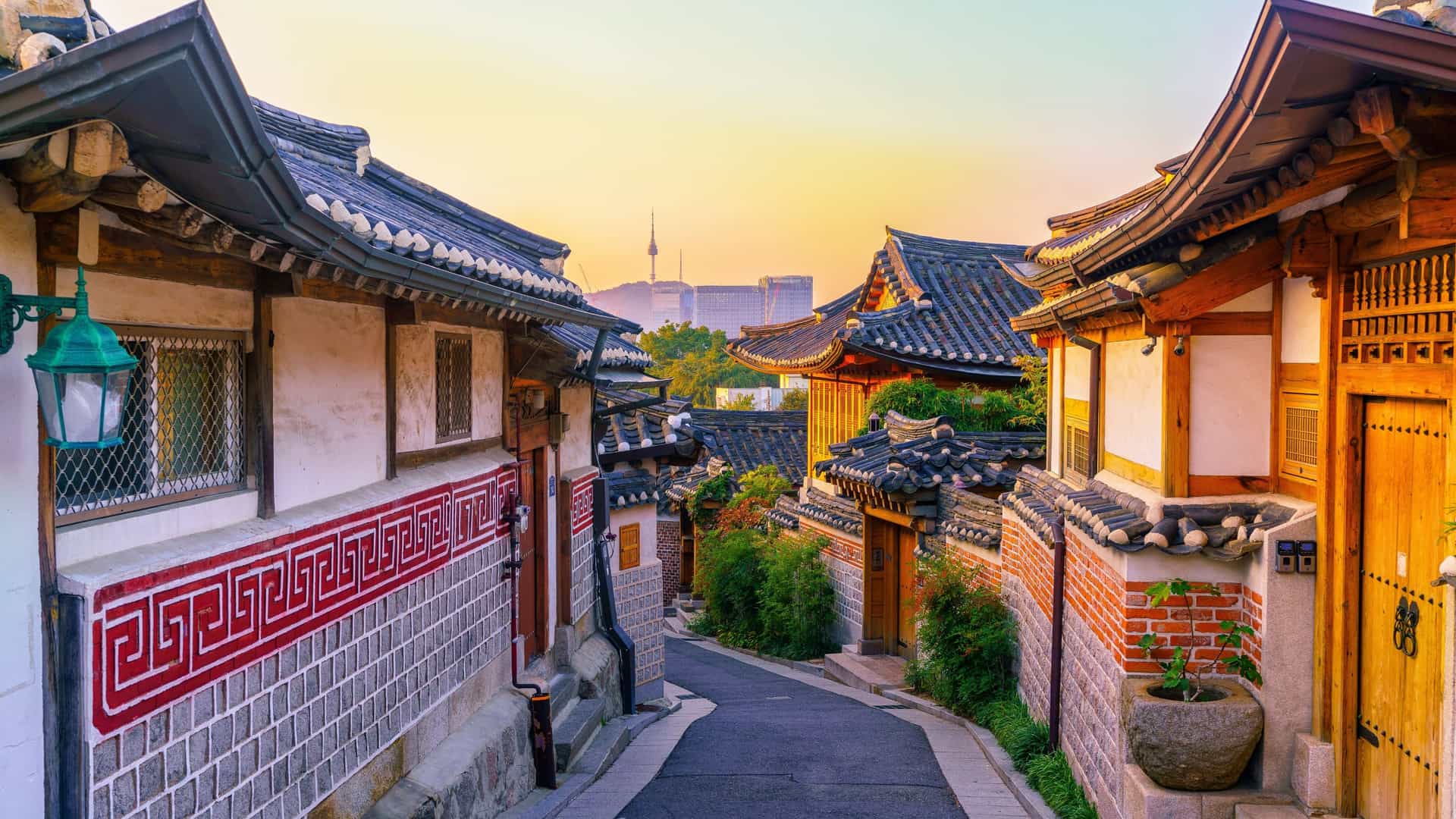 A photo depicting Bukchon Hanok Village in Seoul, South Korea. The image shows a collection of traditional Korean houses, known as hanok, with their characteristic tiled roofs. The village is nestled amongst the modern cityscape of Seoul, often providing a contrast between the historical architecture and contemporary high rise buildings in the distance. The village is a popular tourist destination, showcasing a preserved example of a traditional residential area.