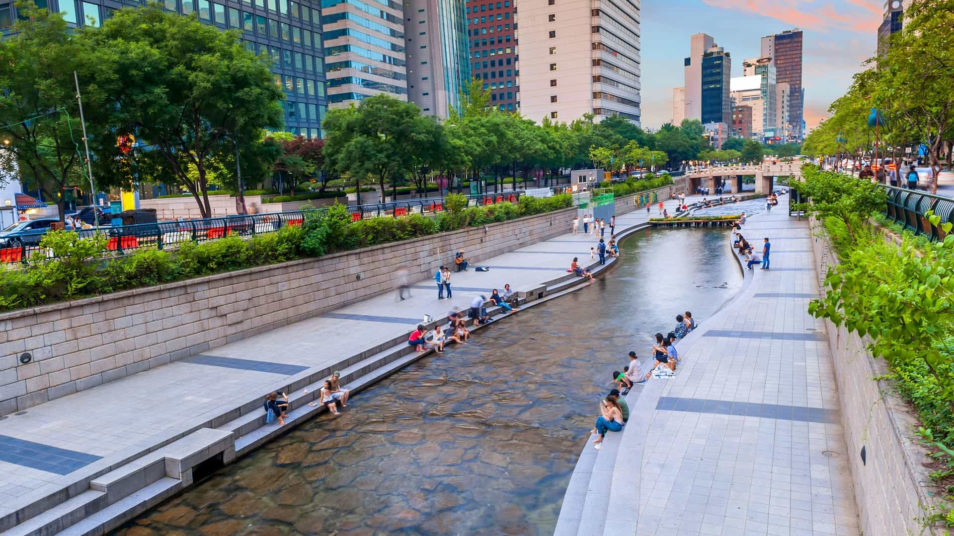 A photograph of the Cheonggyecheon Stream in Seoul, South Korea, showing a view of the modern urban park. The restored stream flows in the center, flanked by concrete walkways and lush greenery, with numerous people scattered along the banks, relaxing and walking. Modern office buildings and city infrastructure line the top of the stream's walls, highlighting the stream's location in the busy downtown area.