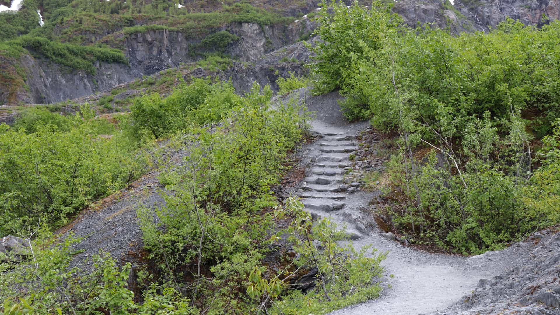 A wooden trail and boardwalk leading through the glacial outwash plain towards the massive, receding ice face of Exit Glacier in Kenai Fjords National Park, Alaska. The foreground features a rugged, rocky landscape and sparse pioneer vegetation, with the steep, snow capped mountains and the deep blue white ice of the glacier filling the background.