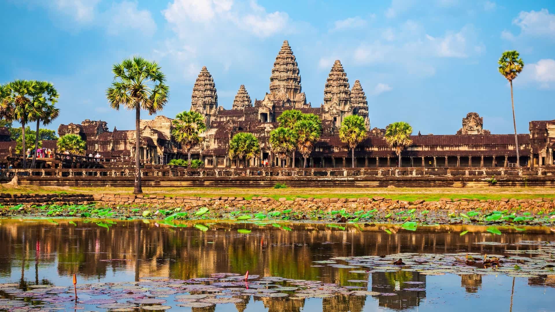 The iconic Angkor Wat temple complex in Siem Reap, Cambodia, featuring its five principal sandstone towers (a quincunx) majestically rising against the sky. The ancient Khmer architecture is reflected in the tranquil water of a foreground pond, surrounded by green foliage.