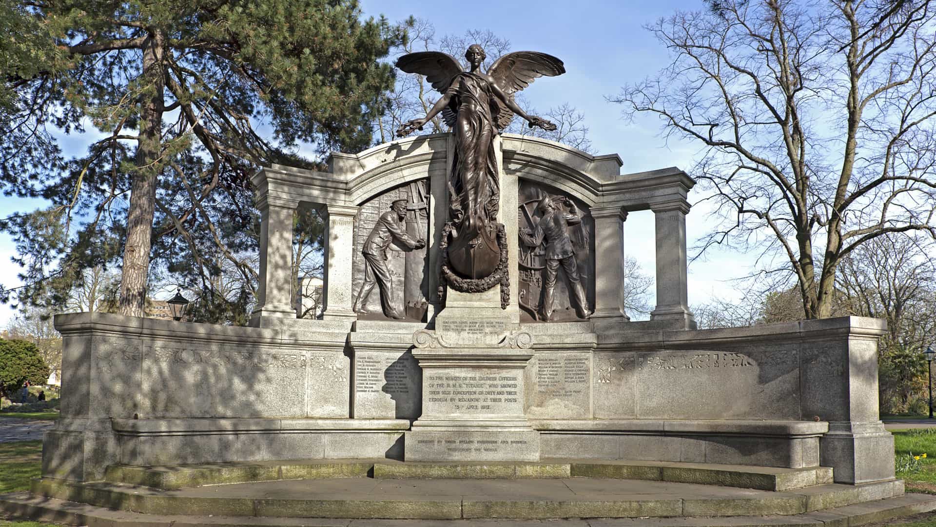 A photo of the Titanic Engineers' Memorial, a Grade II listed monument in Andrews (East) Park, Southampton. The granite and bronze memorial features a central bronze figure of Nike, the Greek Winged Goddess of Victory, standing on the prow of a ship, with bas relief panels on either side depicting the ship's engineers at their posts. The inscription reads: "GREATER LOVE HATH NO MAN THAN THIS. THAT A MAN LAY DOWN HIS LIFE FOR HIS FRIENDS."