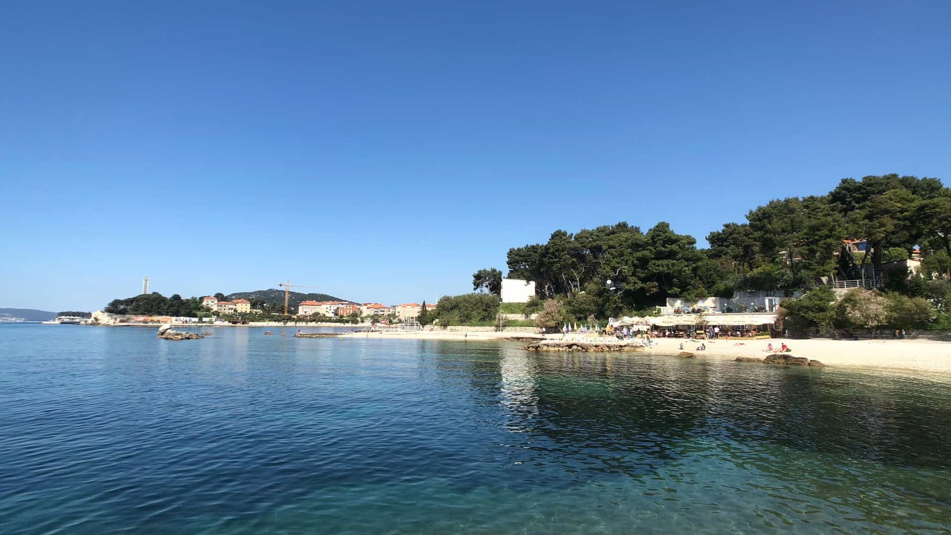 An image of Bacvice Beach in Split, Croatia, showing the popular sandy shore and shallow, clear turquoise waters of the Adriatic Sea. The beach is lined with a concrete promenade, and the background may include a section of the city, beach bars, and people swimming, sunbathing, or playing the traditional Croatian game of picigin.