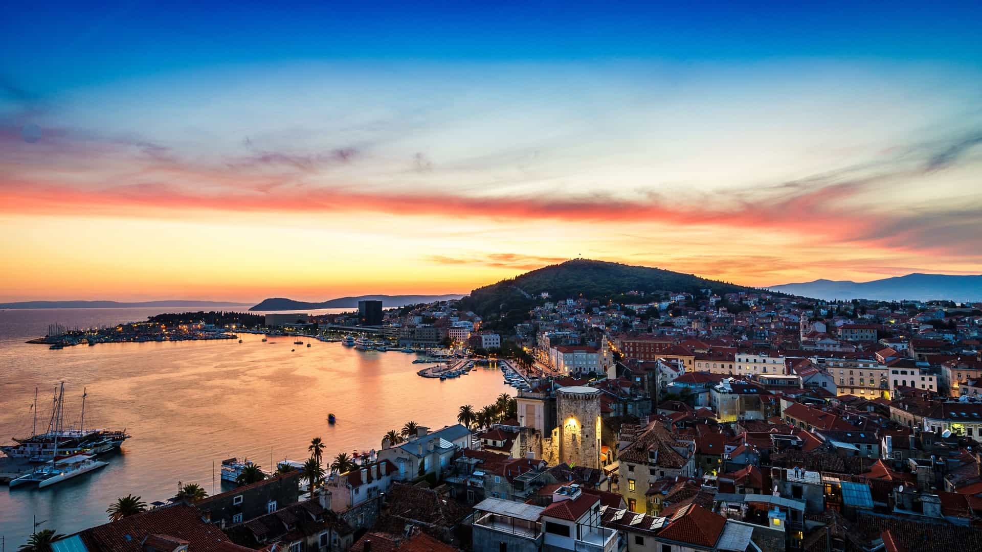 An aerial, panoramic view of the Split waterfront and Marjan Hill in Dalmatia, Croatia, at sunset. The historic city's red tiled rooftops, the palm lined Riva promenade, and the harbor are visible, with the forested Marjan Hill rising prominently in the background against the Adriatic Sea, bathed in the warm, colorful light of dusk.