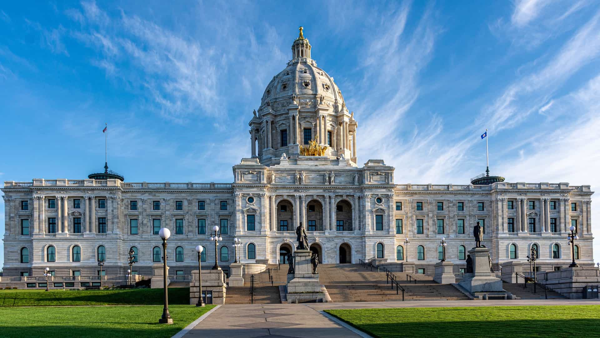The exterior of the Minnesota State Capitol Building in Saint Paul, USA, a grand Beaux Arts structure completed in 1905. The building features a large, unsupported white marble dome, topped by the gold leaf statue grouping known as the Quadriga, titled 'The Progress of the State.' The capitol is viewed from the front lawn under a clear sky, highlighting its classical architecture.