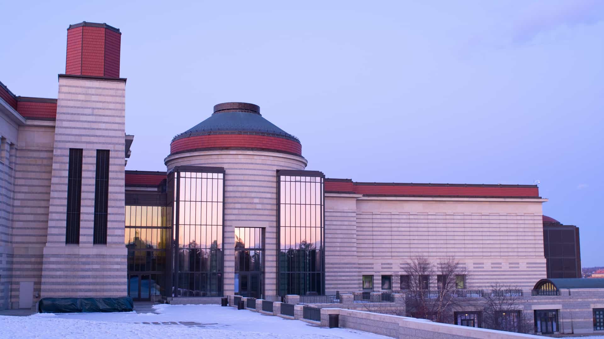 The exterior of the Minnesota History Center building in Saint Paul at dusk. The building, constructed with Minnesota stone and granite, features a large glass atrium and a prominent outdoor terrace. The interior lights are illuminated, contrasting with the dark blue evening sky.