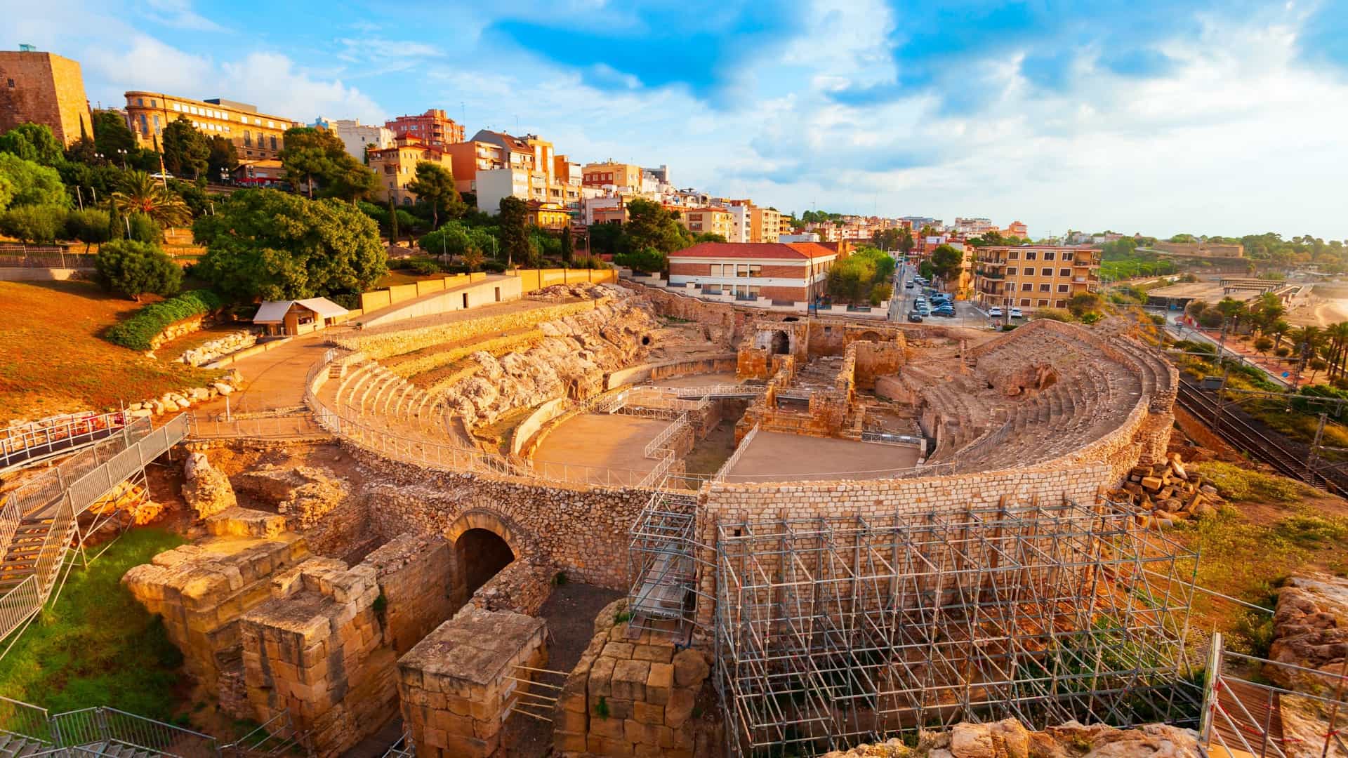 An aerial, panoramic view of the oval Roman Amphitheatre ruins in Tarragona, Spain, a UNESCO World Heritage site, set dramatically on the coast of the Mediterranean Sea. The ancient stone arena sits near the bottom center, framed by green trees, with a strip of sandy beach and the vast, deep blue sea stretching to the horizon. The modern city of Tarragona is visible along the top and right side of the photo, contrasting the historical ruins.