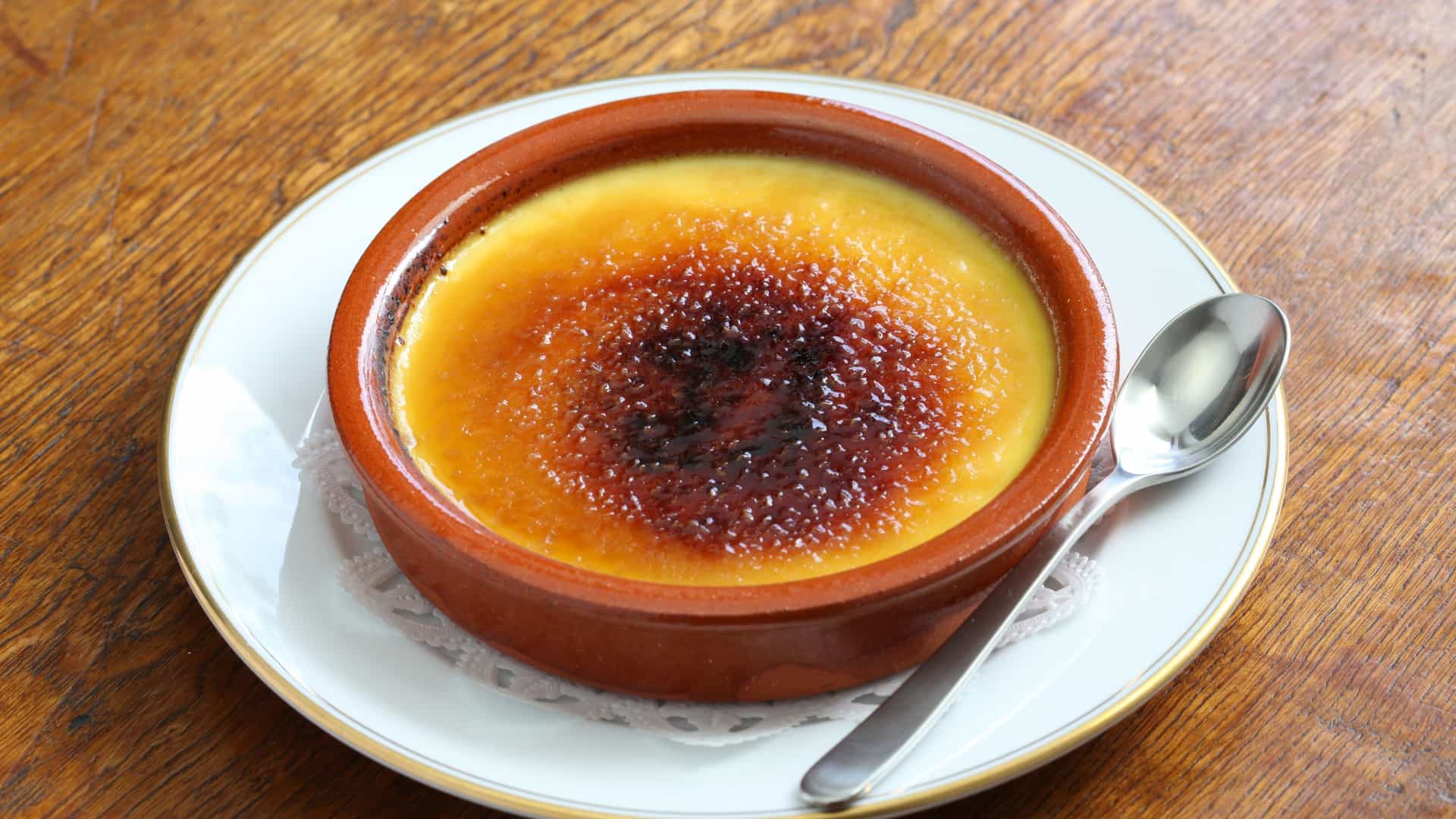 A close up of a serving of Crema Catalana, a Spanish dessert. The creamy yellow custard is presented in a white ramekin, topped with a thin, brittle layer of caramelized sugar that has been cracked in several places, revealing the smooth custard beneath. The dessert is garnished with a single cinnamon stick.