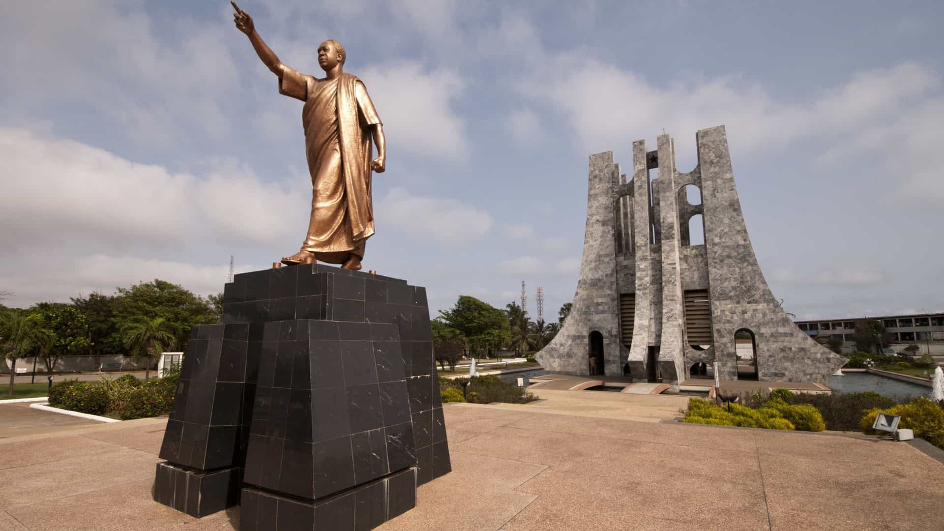 A view of the Kwame Nkrumah Memorial Park in Accra, Ghana. The modernist, granite and Italian marble mausoleum, which is shaped like an upside down sword—a symbol of peace—and capped with a black star, stands as the park's centerpiece. In the foreground, the bronze statue of Ghana's first president, Osagyefo Dr. Kwame Nkrumah, stands in a commanding pose, often with his hand raised or outstretched. The monument is surrounded by a reflecting pool with fountains and is set against a backdrop of lush green grass and trees.