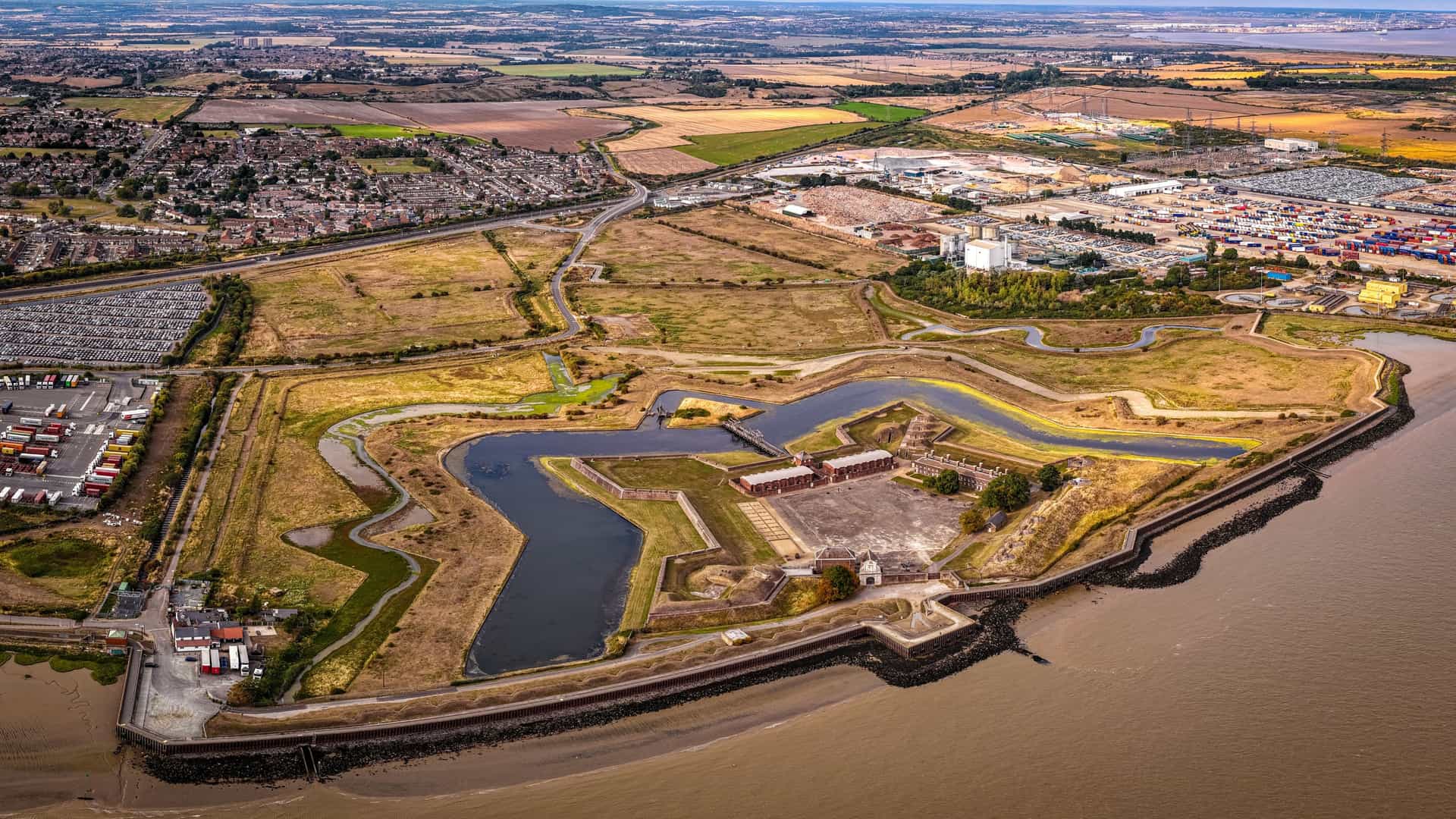 An aerial drone shot of the historic Tilbury Fort in Essex, UK. The image captures the fort's distinctive 17th century, star shaped defensive design, including its angular brick bastions, surrounding water filled moats, and outworks, all situated on the north bank of the River Thames. The surrounding landscape includes marshland and the wide river.