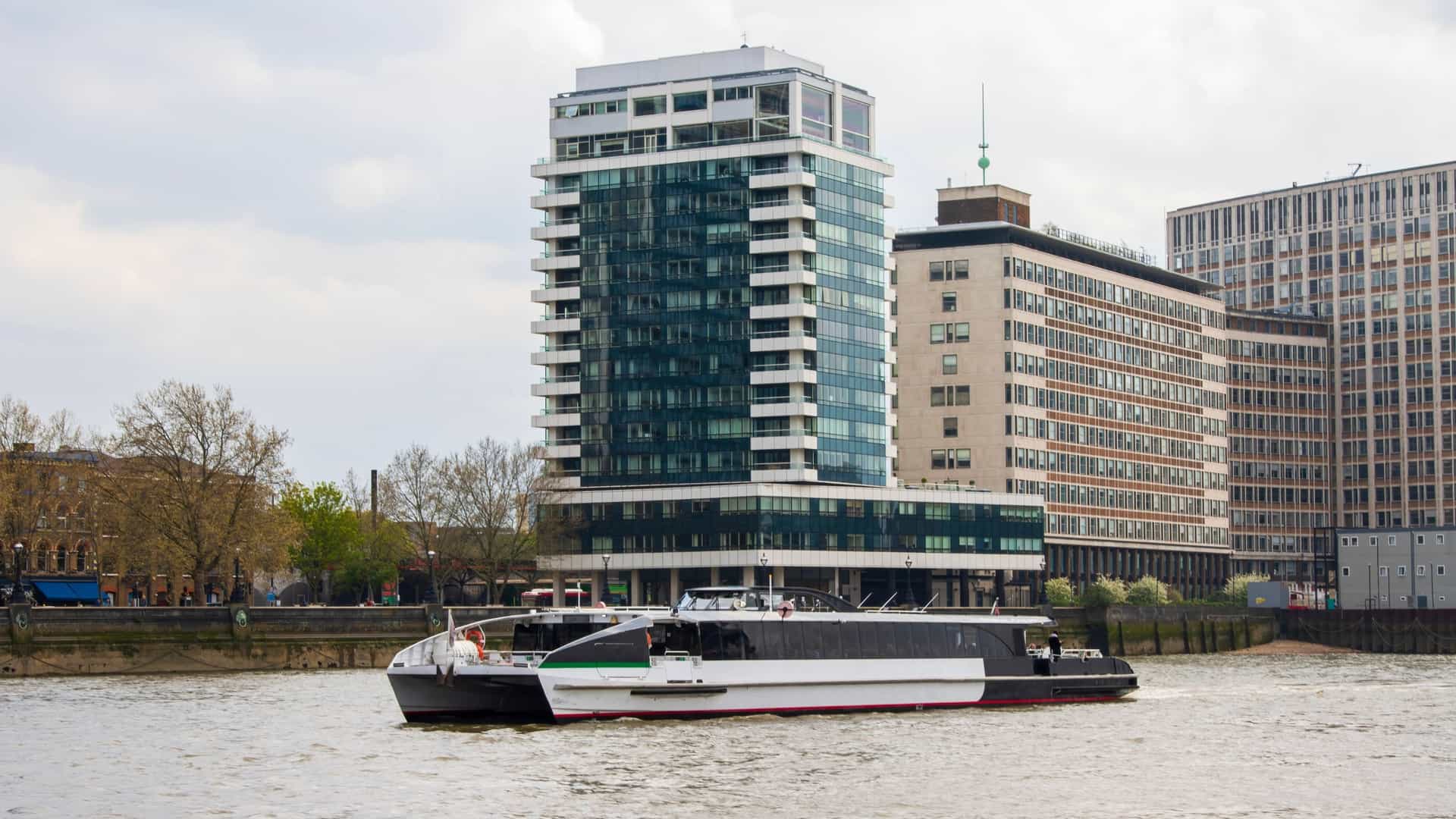 A river bus service, branded as Uber Boat by Thames Clippers, is traveling along the River Thames in central London on a clear day. The sleek, high speed catamaran is seen from the side, moving past a pier or a section of the riverbank. This river bus provides both commuter and tourist services, offering a unique and scenic way to traverse the city.