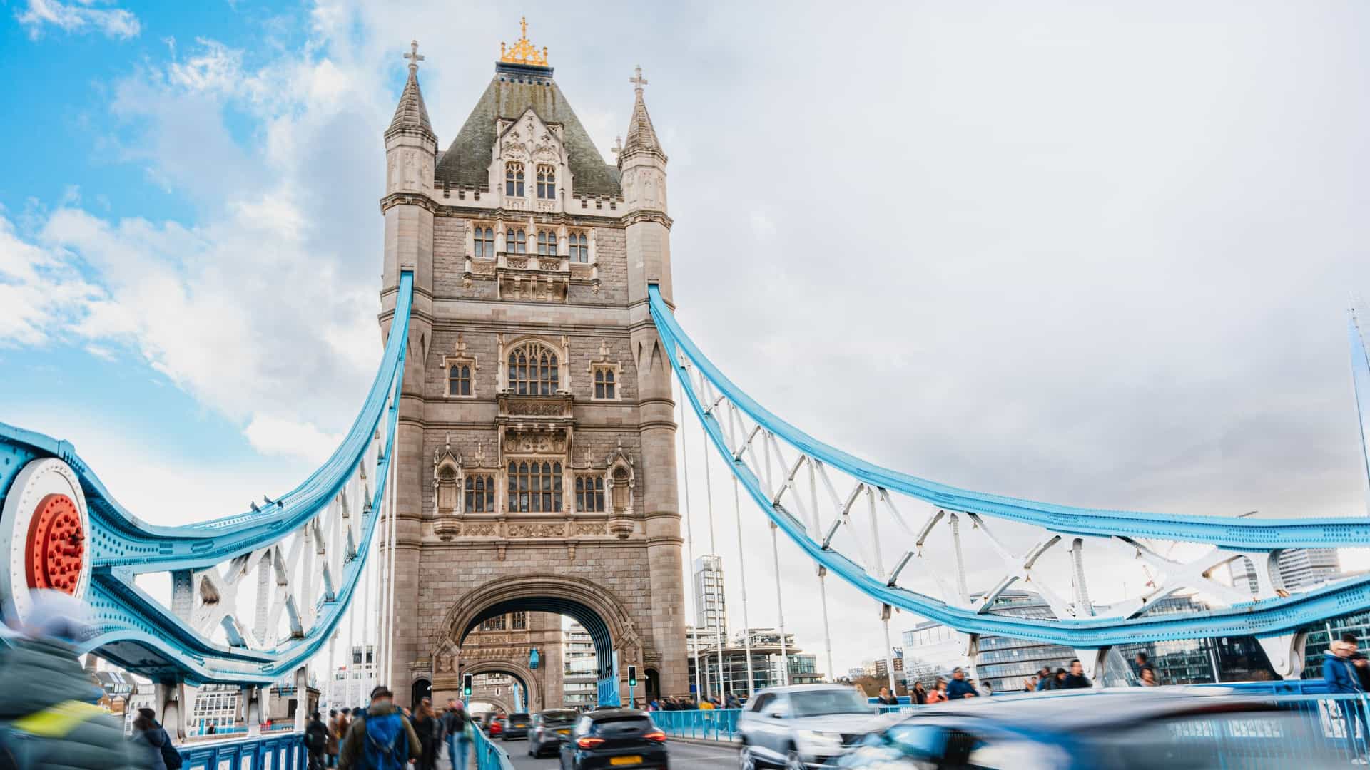 The image depicts Tower Bridge in London, likely showing the lower road deck with vehicles and people moving across it. The bascules (movable sections of the road) are down, allowing traffic to flow. The iconic Neo Gothic towers and the high level walkways connecting them are visible. The River Thames flows beneath the bridge, and the scene is a busy representation of one of London's most famous landmarks acting as a vital modern traffic route.