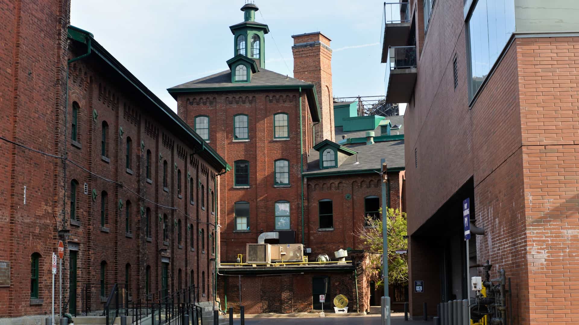 A view of Toronto's historic Distillery District, showcasing its characteristic Victorian era red brick industrial buildings lining a pedestrian only, cobblestone street. The scene features a blend of historical architecture now occupied by boutiques, cafes, and restaurants, creating an old world atmosphere.