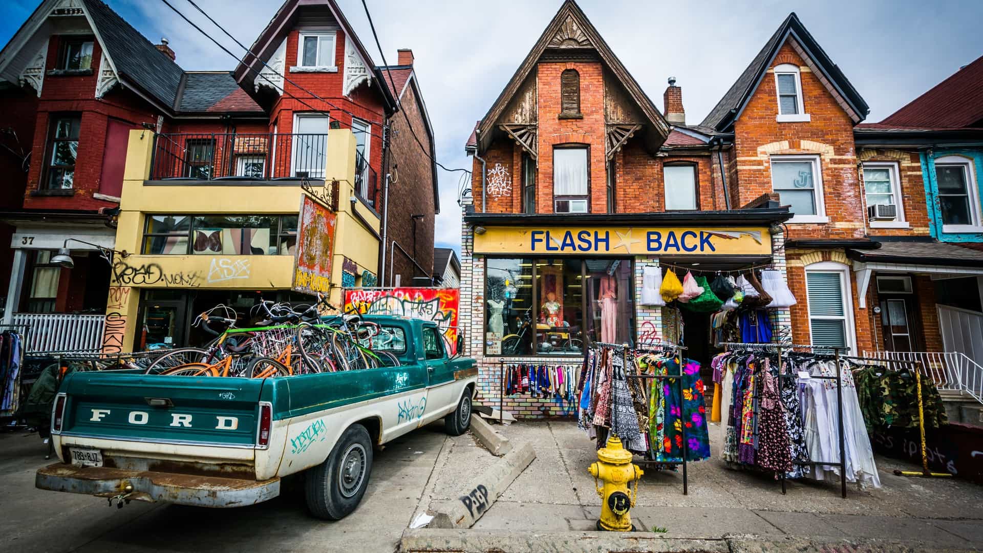 A vibrant street scene on Kensington Avenue in Toronto's eclectic Kensington Market. The view features a colorful mix of low rise buildings and shops with street level retail, a characteristic feature of this bohemian and multicultural neighborhood.