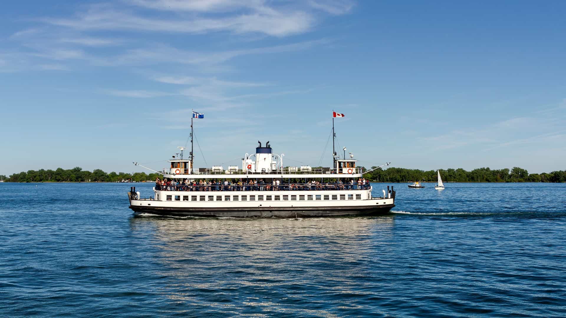 A passenger ferry boat, part of the fleet transporting visitors to the Toronto Islands, is cruising across Lake Ontario, leaving Centre Island with the distinctive Toronto skyline, including the CN Tower, visible in the distance.