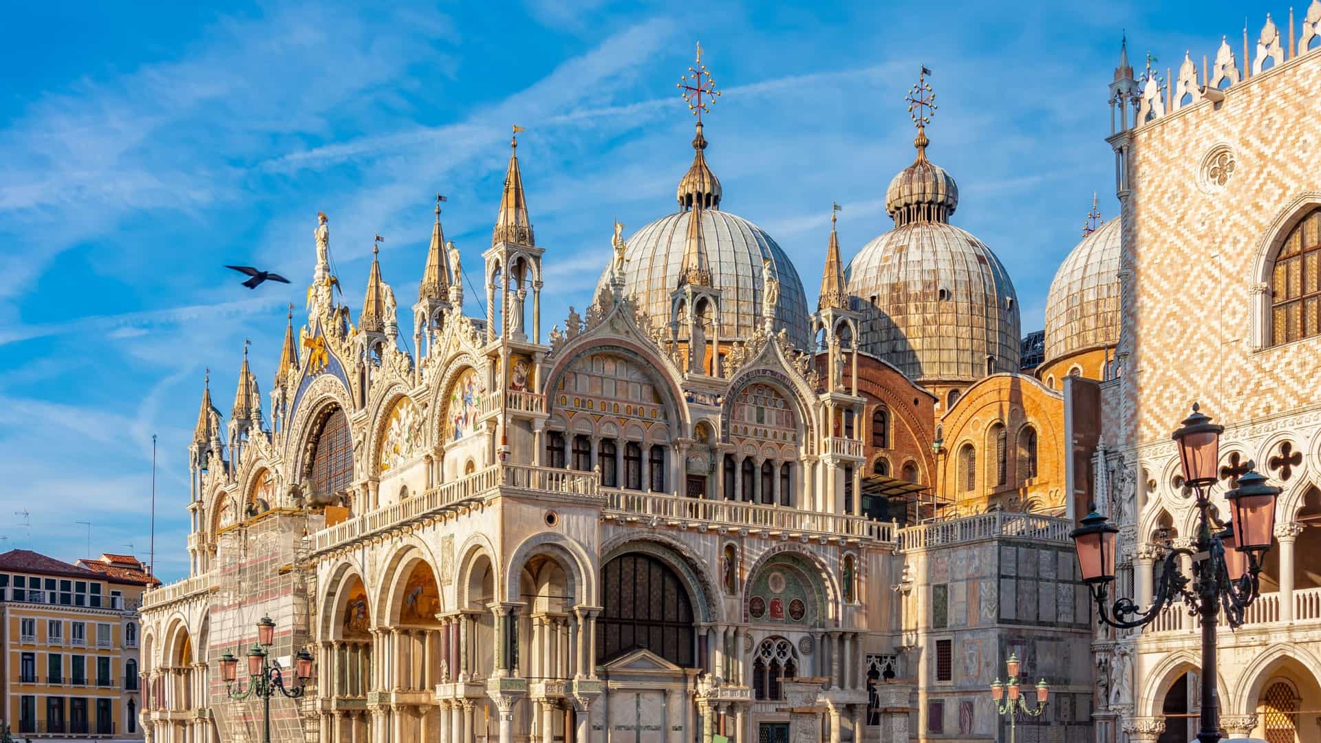 The ornate, Italo Byzantine facade of Saint Mark's Basilica (Basilica di San Marco) in Venice, Italy, showcasing its multiple golden tiled domes, intricate stone carvings, and the five deep arches of the main entrance, topped with statues and mosaics. The central portal features the famous Quadriga (four bronze horses) on the terrace above.