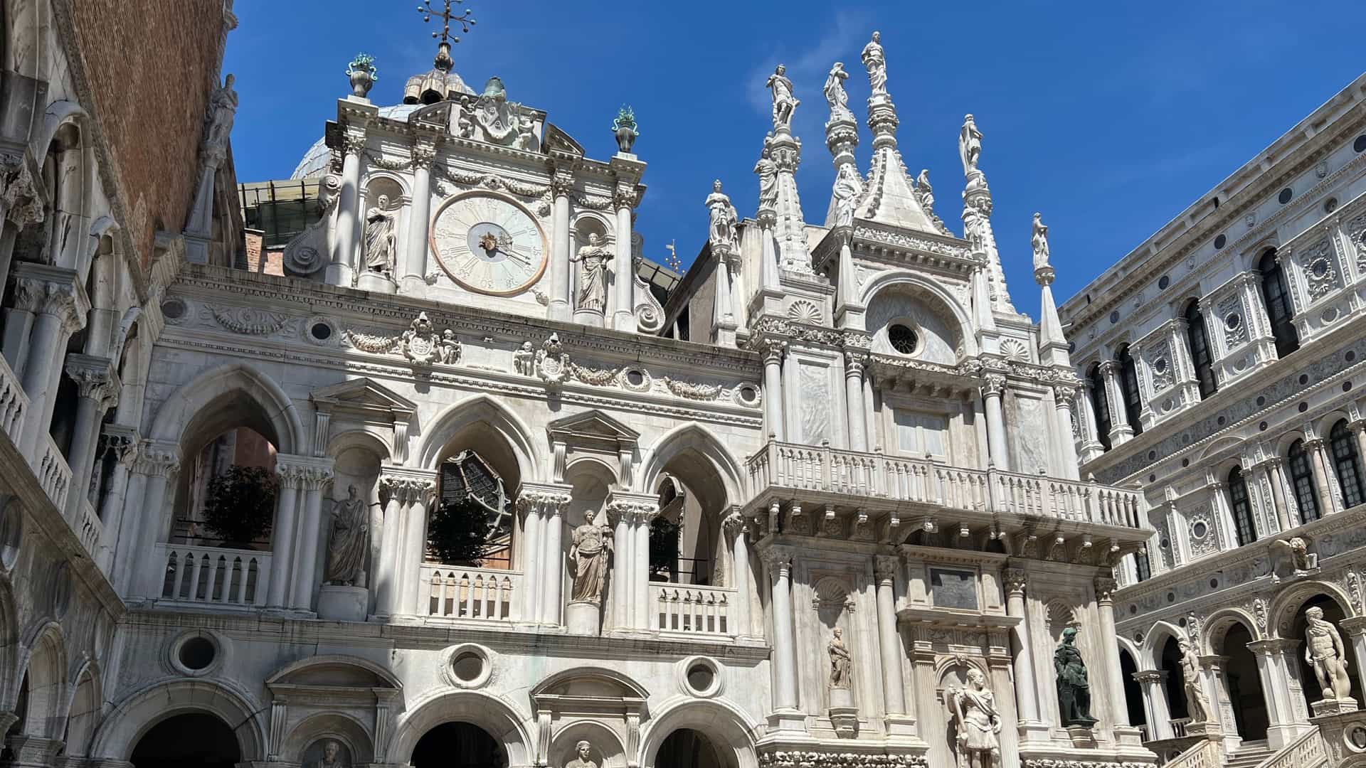 The Doge's Palace (Palazzo Ducale) in Venice, Italy, a masterpiece of Venetian Gothic architecture. The facade features a striking upper story patterned with light pink and white diamond shaped stone, resting upon two levels of open loggias. The lower colonnade consists of a continuous arcade of delicate white Istrian stone columns with ornate capitals, standing along the water's edge.