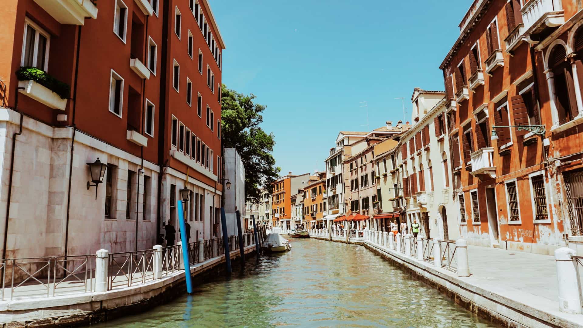 A sun drenched view of a narrow Venetian canal. Historic buildings with aged, colored plaster walls rise on both sides, their reflections visible in the still, deep green water. A small, arched stone footbridge crosses the canal, and a few gondolas or smaller boats are visible either moored or passing underneath.