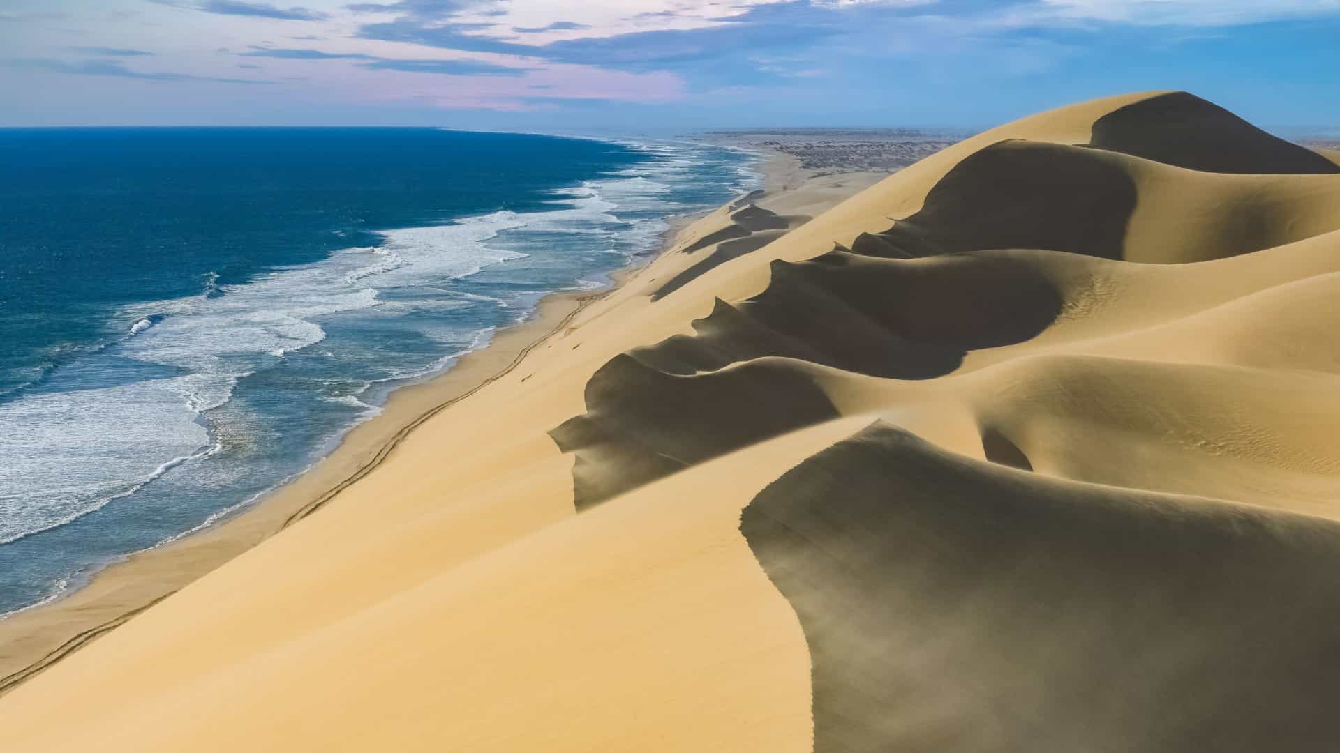 A dramatic landscape photo of the Namib Desert in Namibia, showing huge, sweeping yellow orange sand dunes with sharp, defined ridges and deep shadows, stretching out under a clear blue sky.