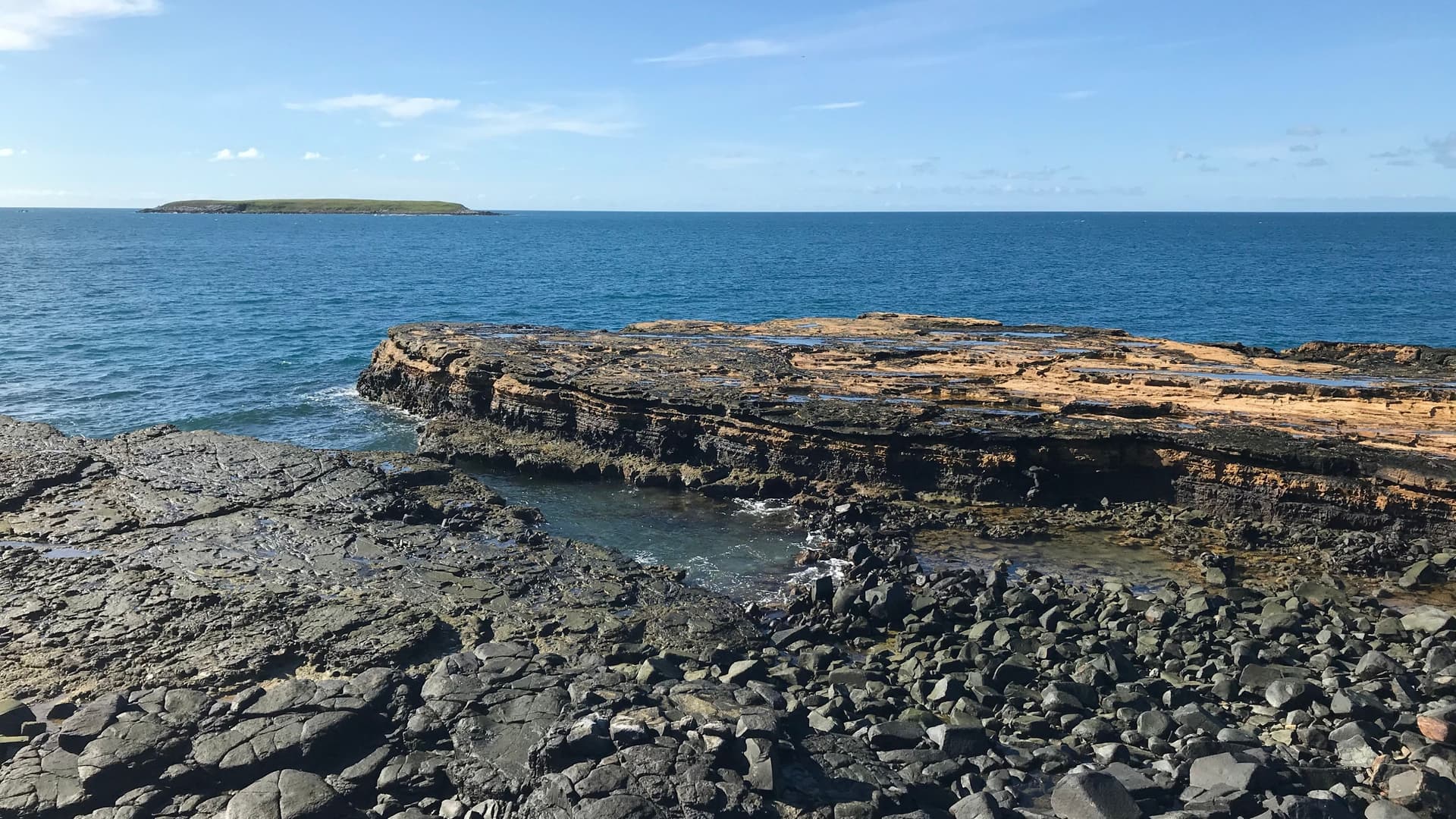 A wide-angle view of the volcanic rock formations and rugged coastline of the Abrolhos Archipelago in Brazil, featuring layered sediment shelves and tide pools overlooking the blue South Atlantic Ocean with a small island visible on the horizon.