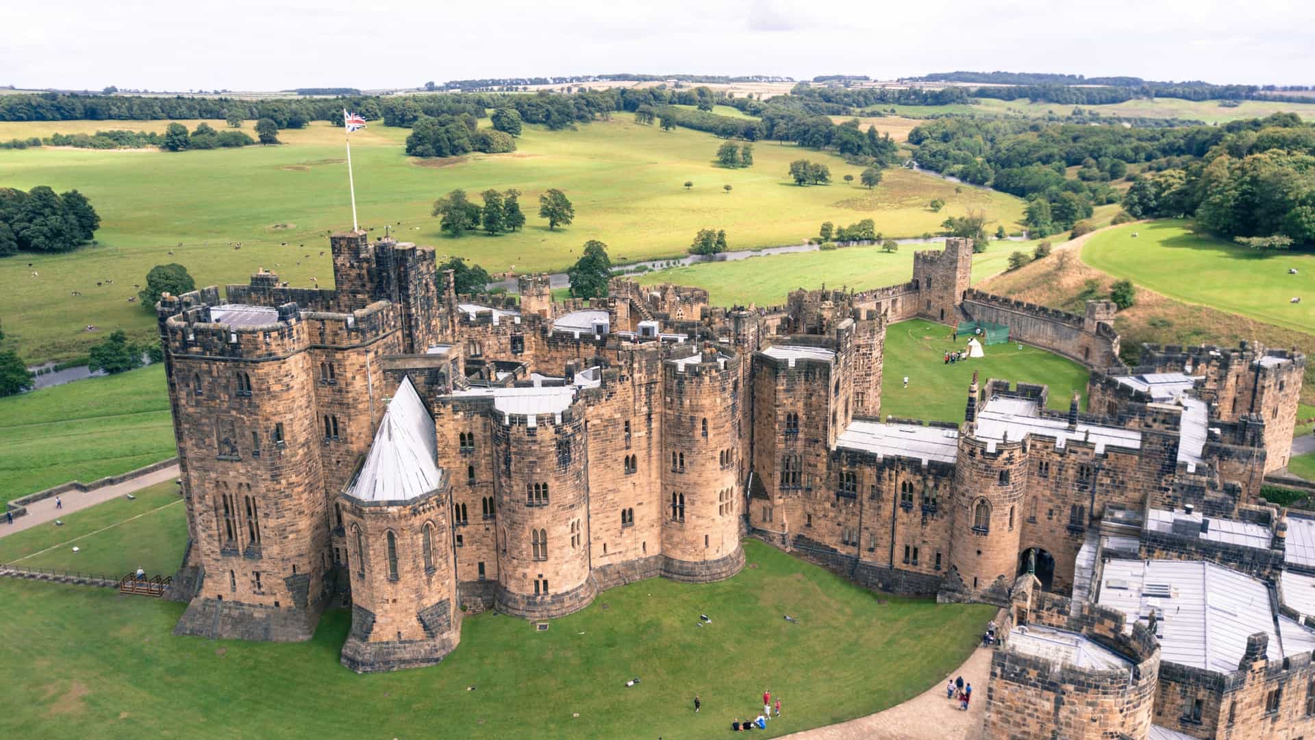 An aerial view of the historic Alnwick Castle in Northumberland, England, showcasing its massive stone walls, medieval battlements, and central courtyard, surrounded by lush green landscapes and the rolling hills of the English countryside.